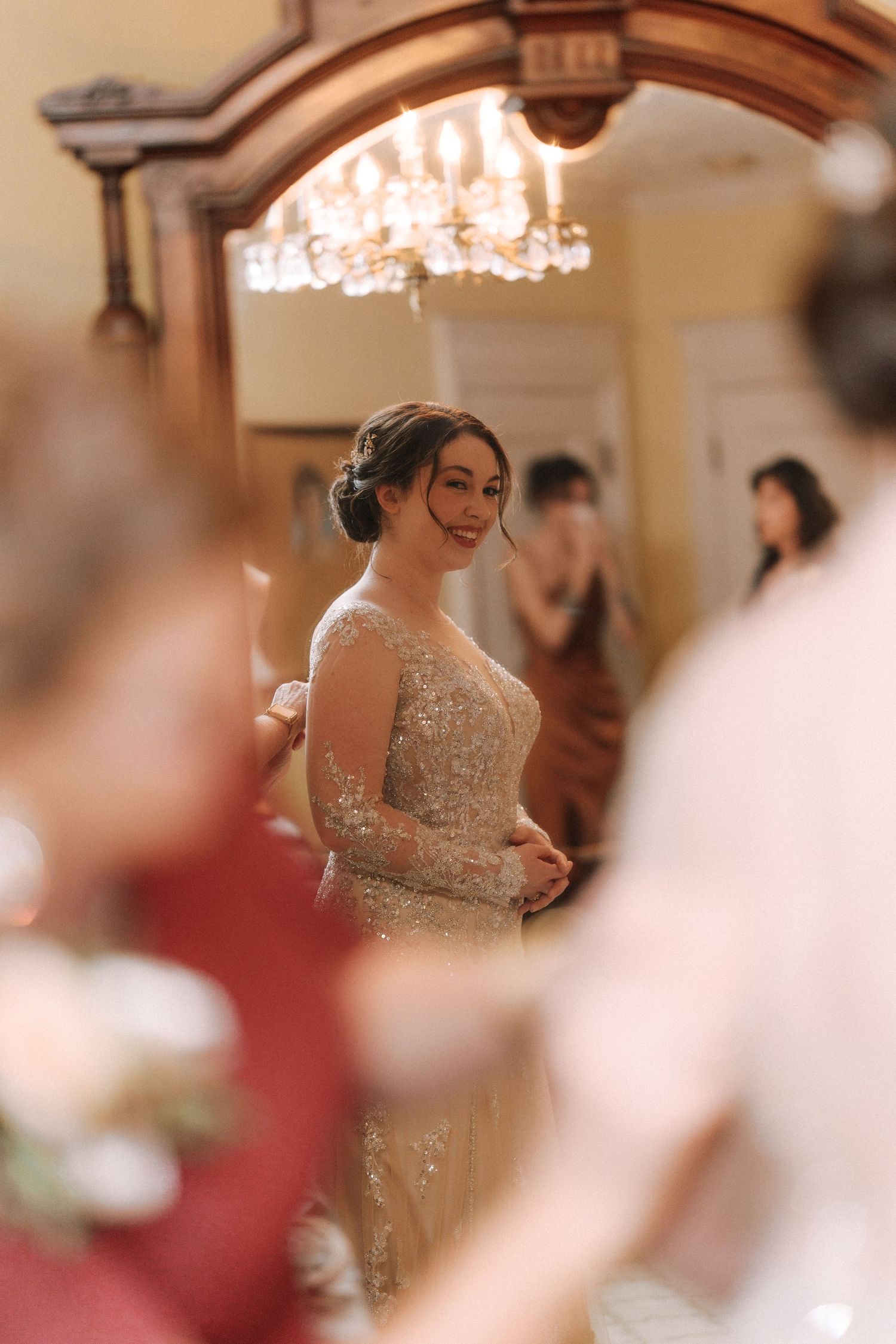 Elegant bride in beaded gown smiling in ornate room with chandelier reflected in antique mirror.