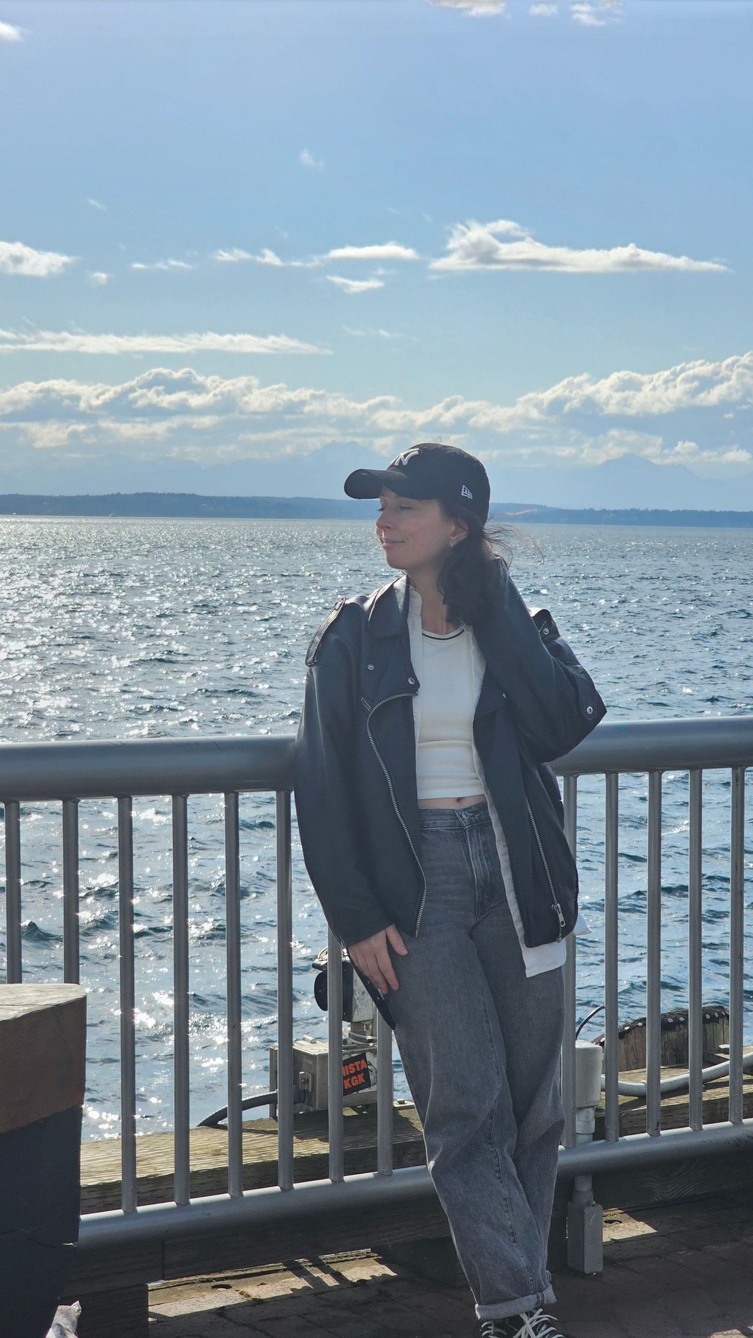 Young woman in black leather jacket and cap posing by waterfront railing on a sunny day with ocean view.