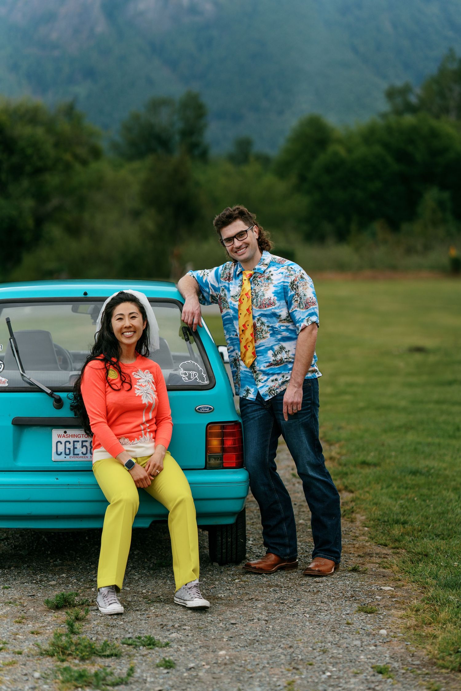 Couple posing by a teal vintage car, woman in yellow pants and coral top, man in floral shirt.