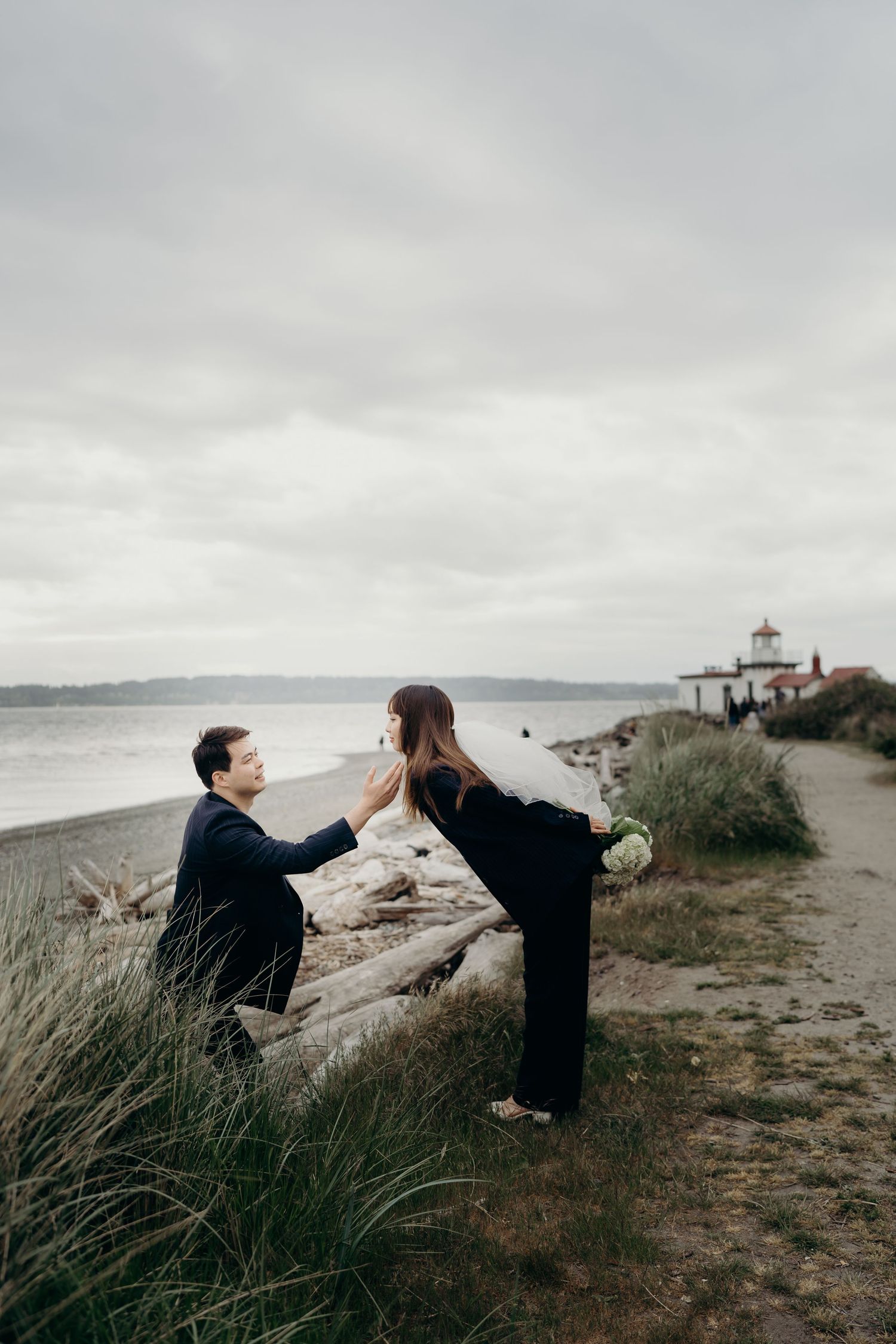Man on one knee proposing to woman on driftwood near beach with lighthouse in background.