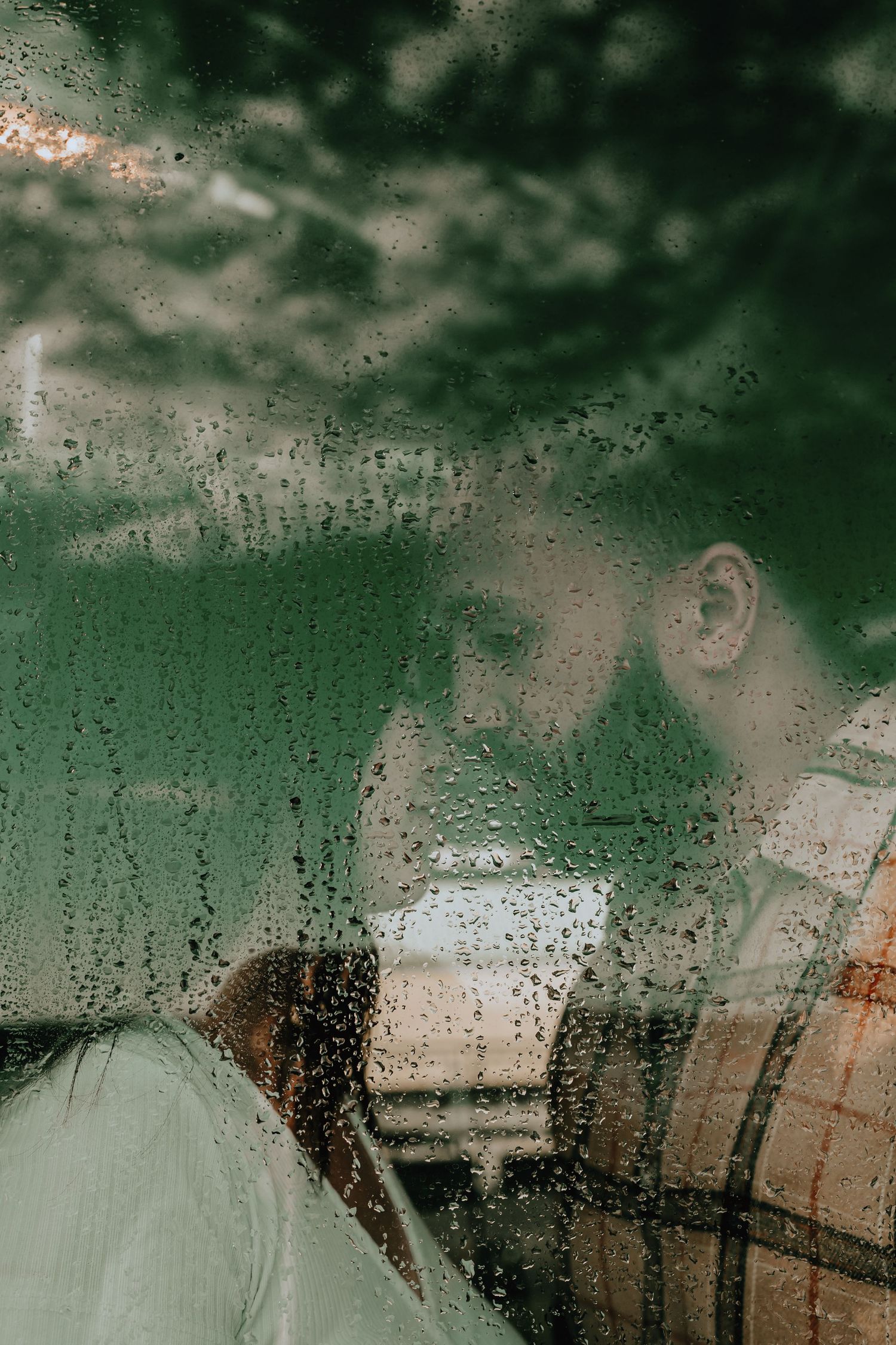 Romantic couple sharing a kiss viewed through a rain-covered window with green foliage behind.