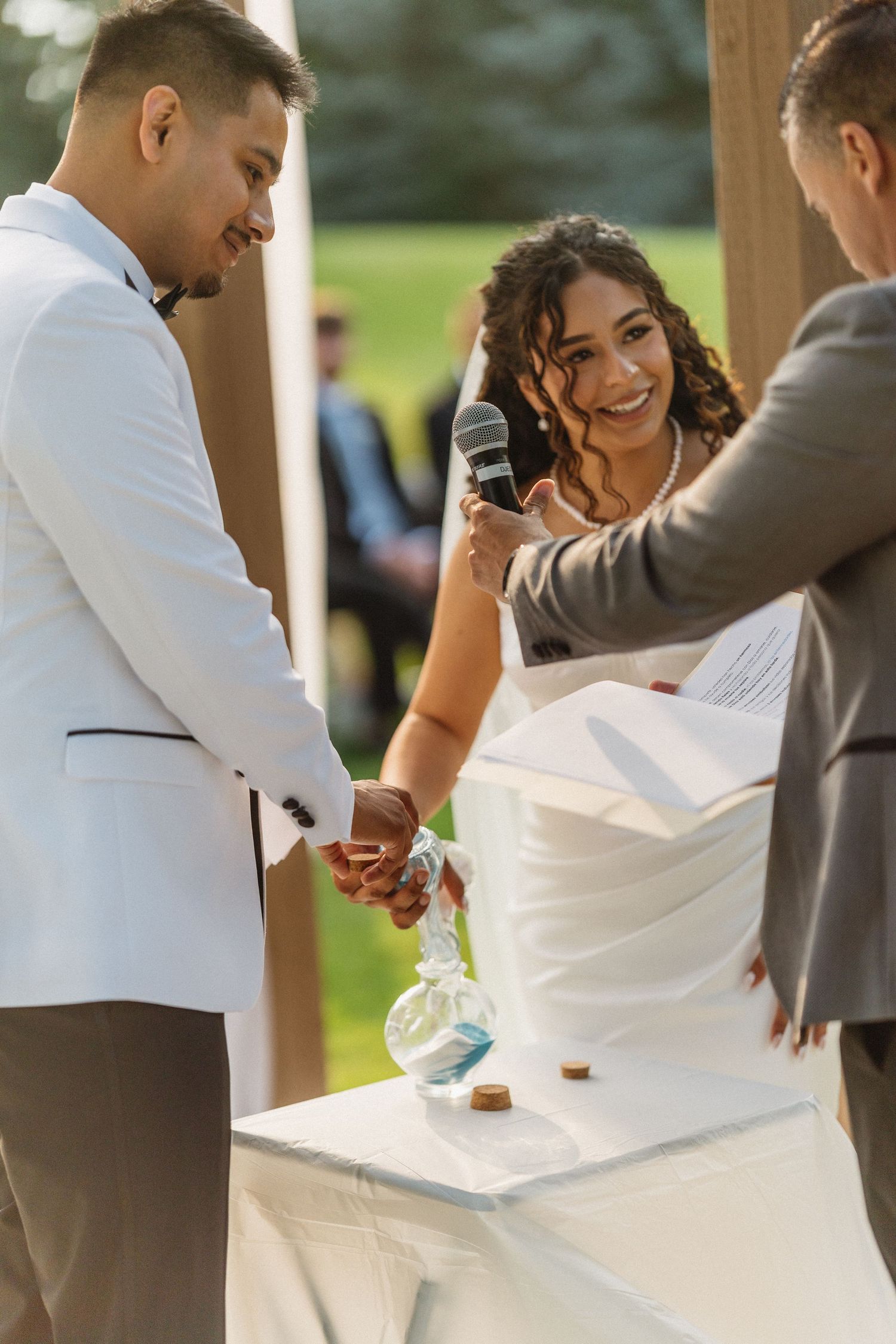 Smiling bride holds groom's hands during outdoor wedding ceremony with officiant.