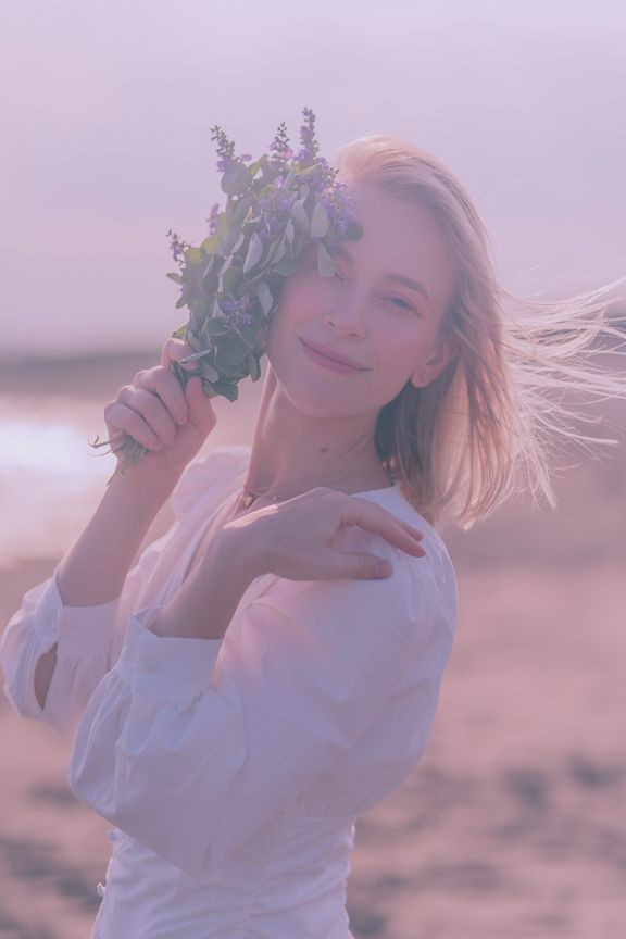 Smiling blonde woman in white blouse holding purple wildflowers at beach during sunset.