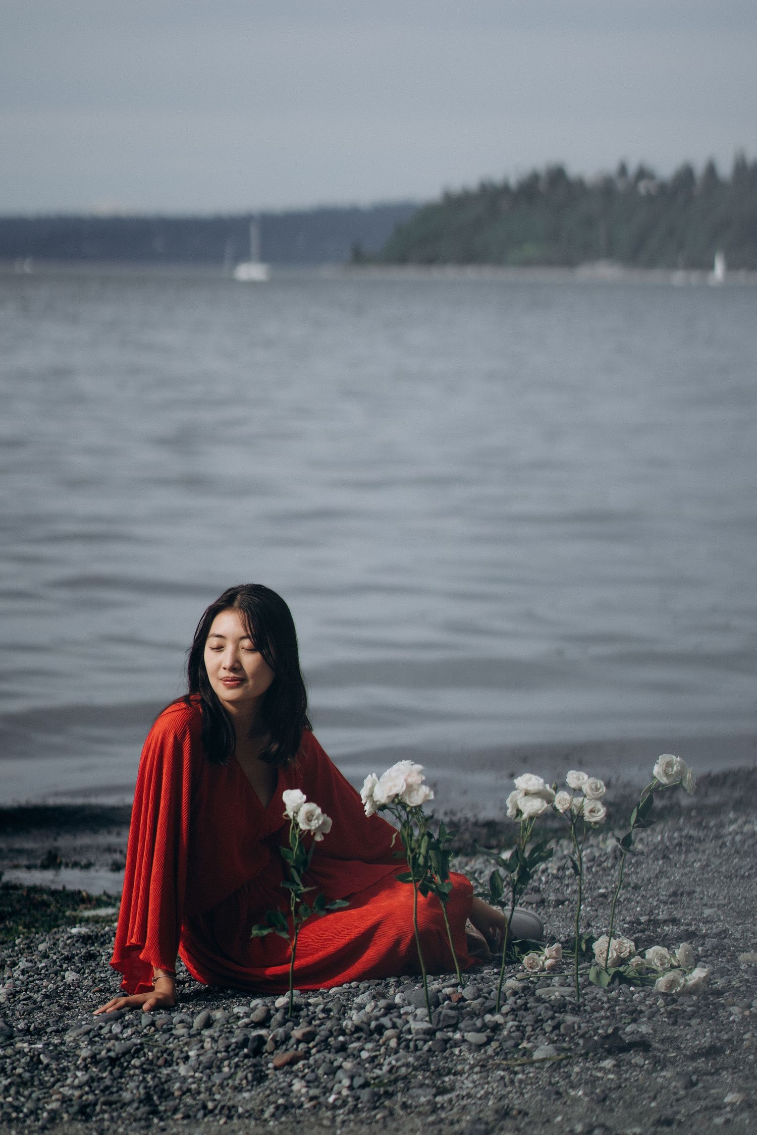 Woman in red dress sitting on a pebble beach beside white flowers near water.
