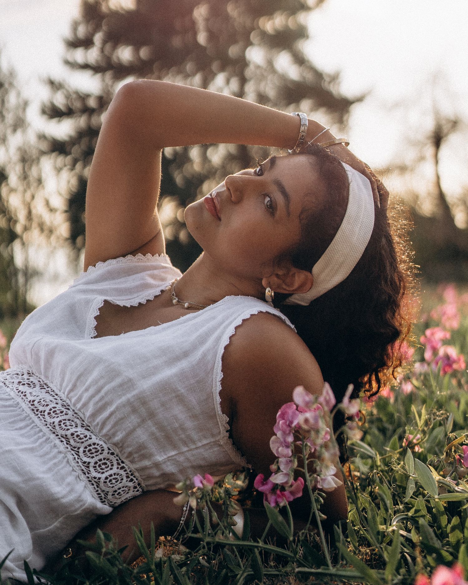 Young woman in white lace dress resting among pink wildflowers in golden hour sunlight with headband.