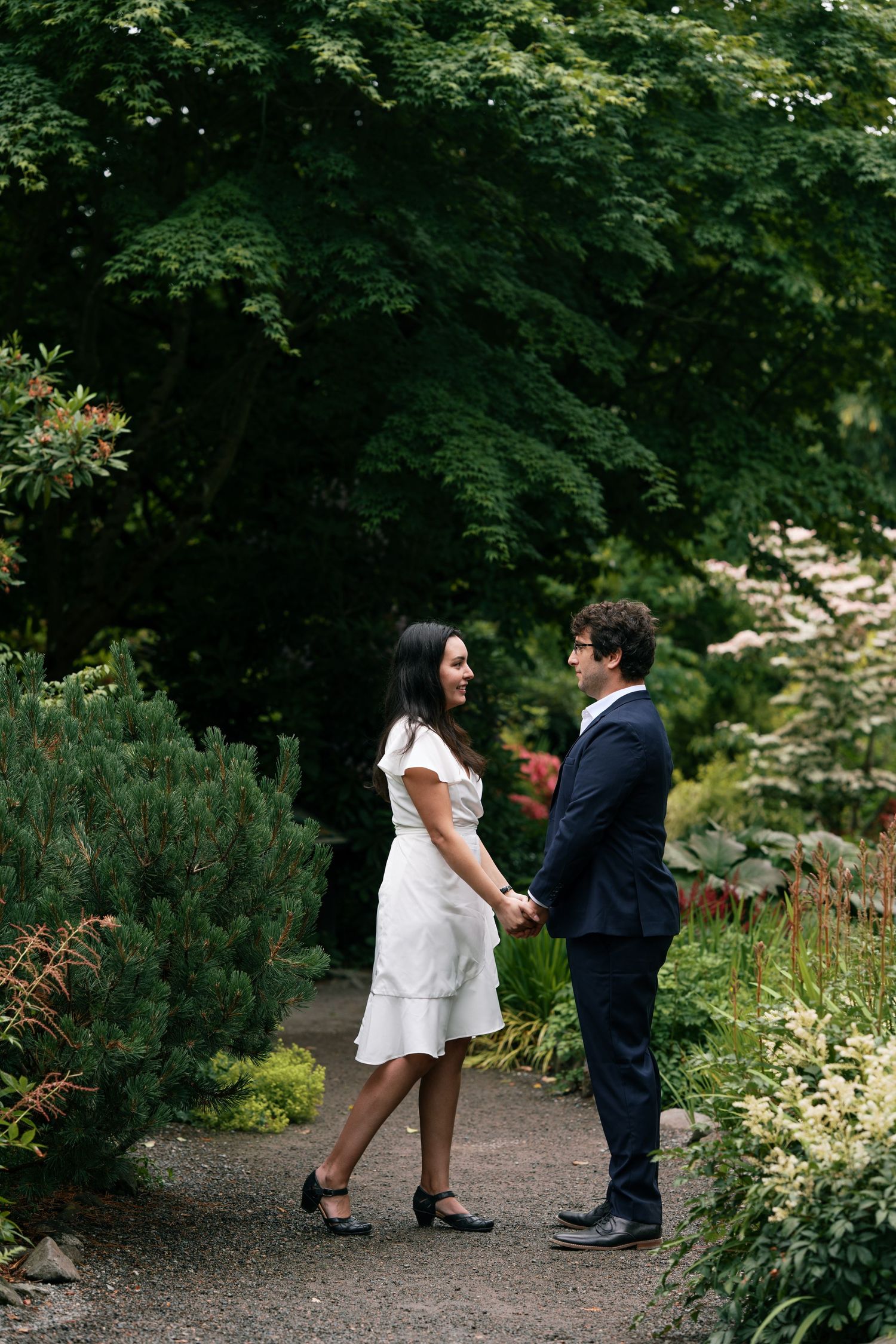 Couple holding hands on a garden path, woman in white dress and man in navy suit surrounded by lush greenery.