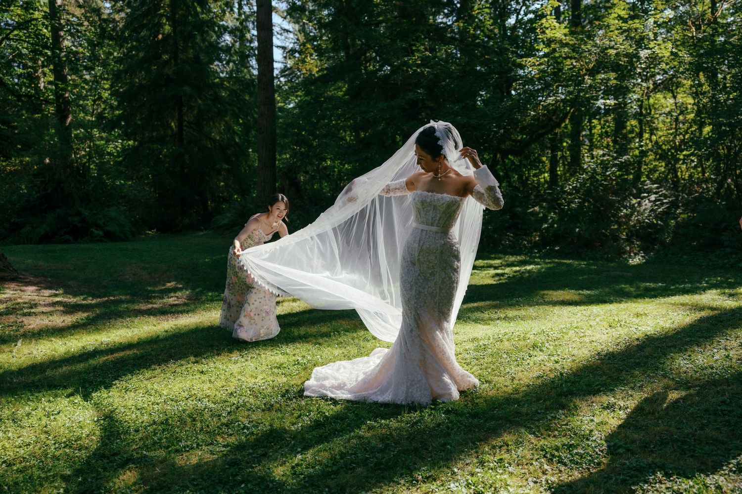 Bridesmaid adjusts bride's flowing cathedral veil on sunlit green lawn in forest.