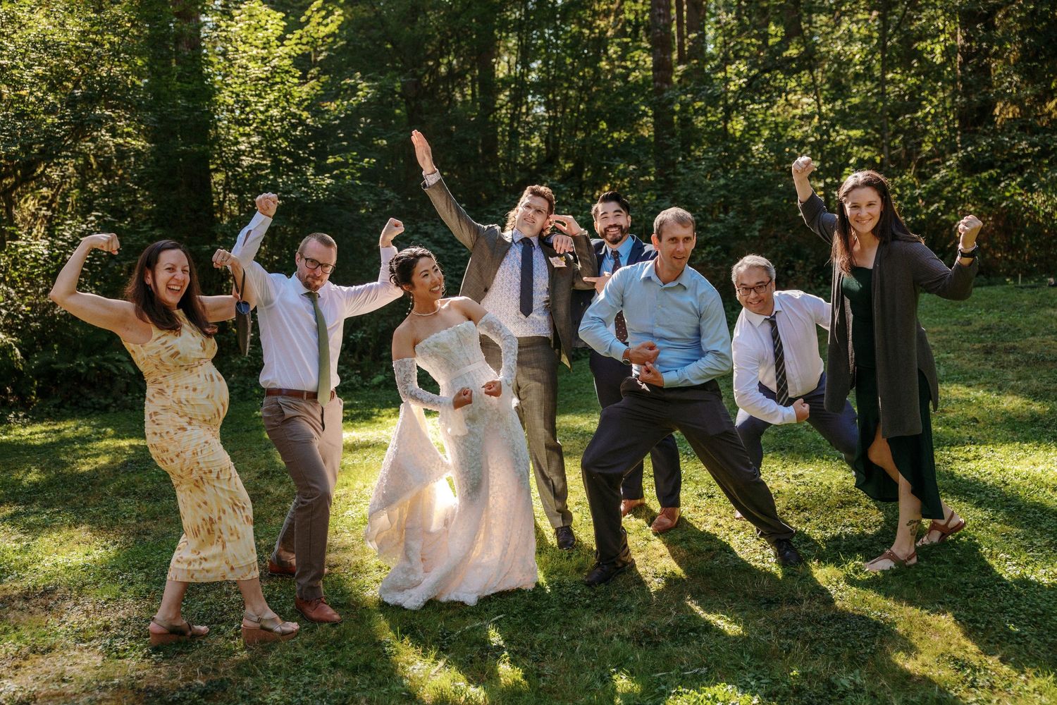 Joyful wedding party celebrates outdoors in a lush green forest setting, with bride in white gown raising arms.