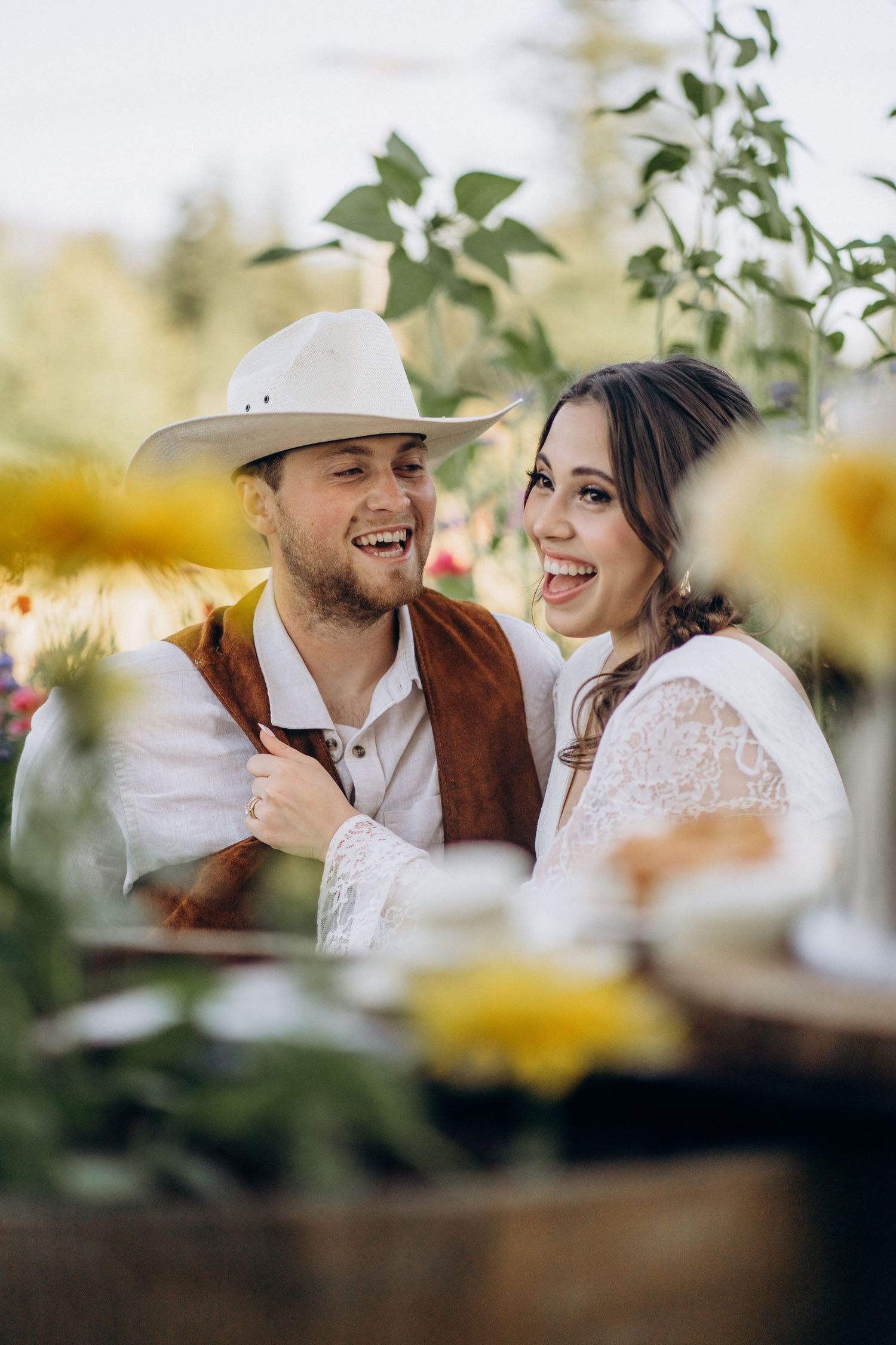 Laughing bride and groom at an outdoor rustic wedding celebration, groom in white cowboy hat and brown suede vest.