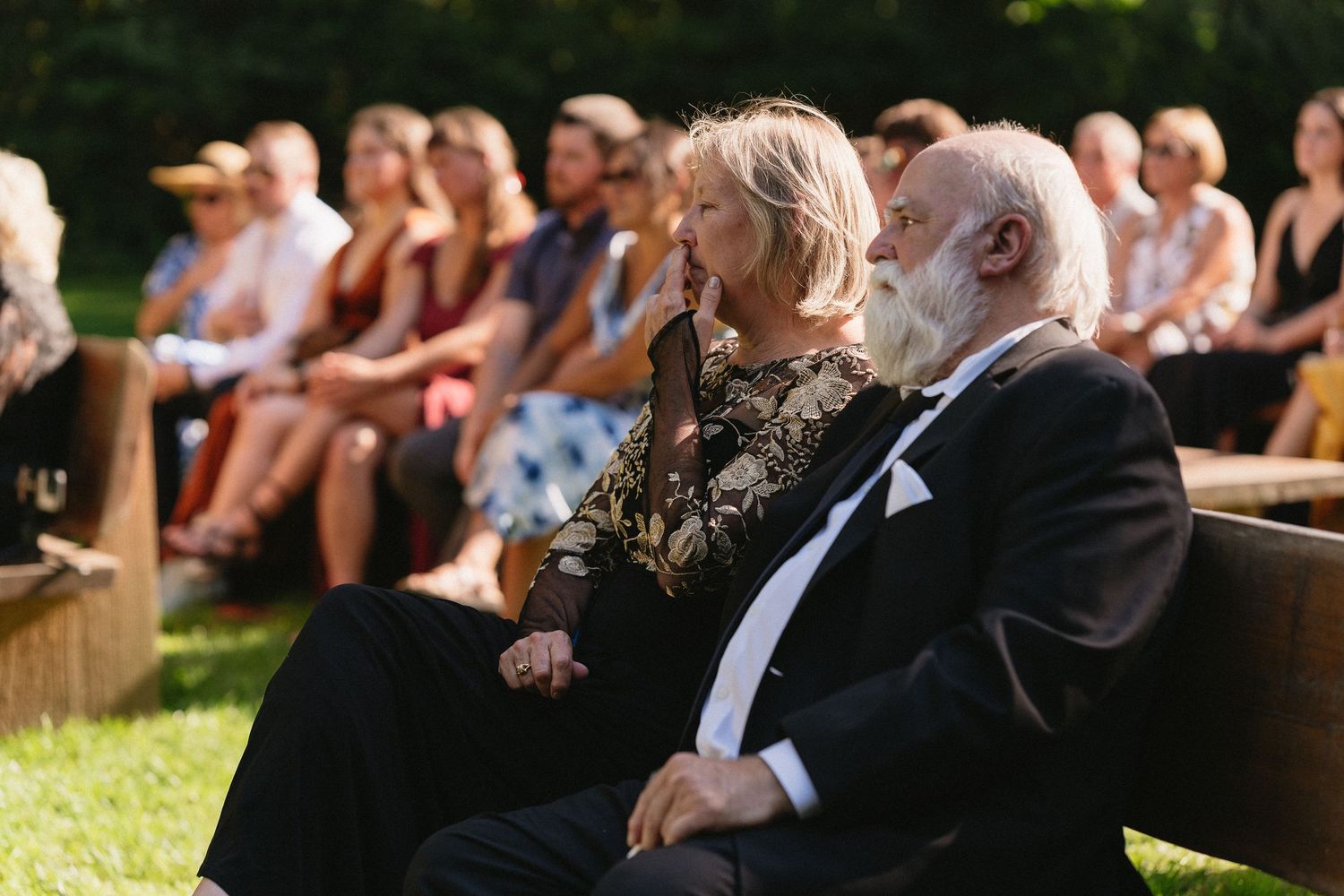 Elderly wedding guests seated outdoors watching ceremony, woman in floral dress beside bearded man.