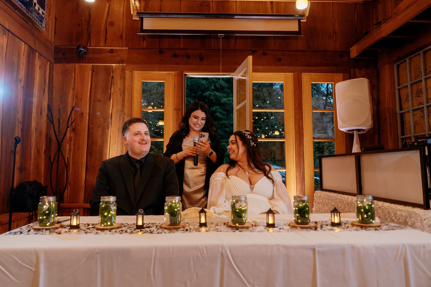 Wedding guest shows phone to smiling bride and groom at decorated reception head table.