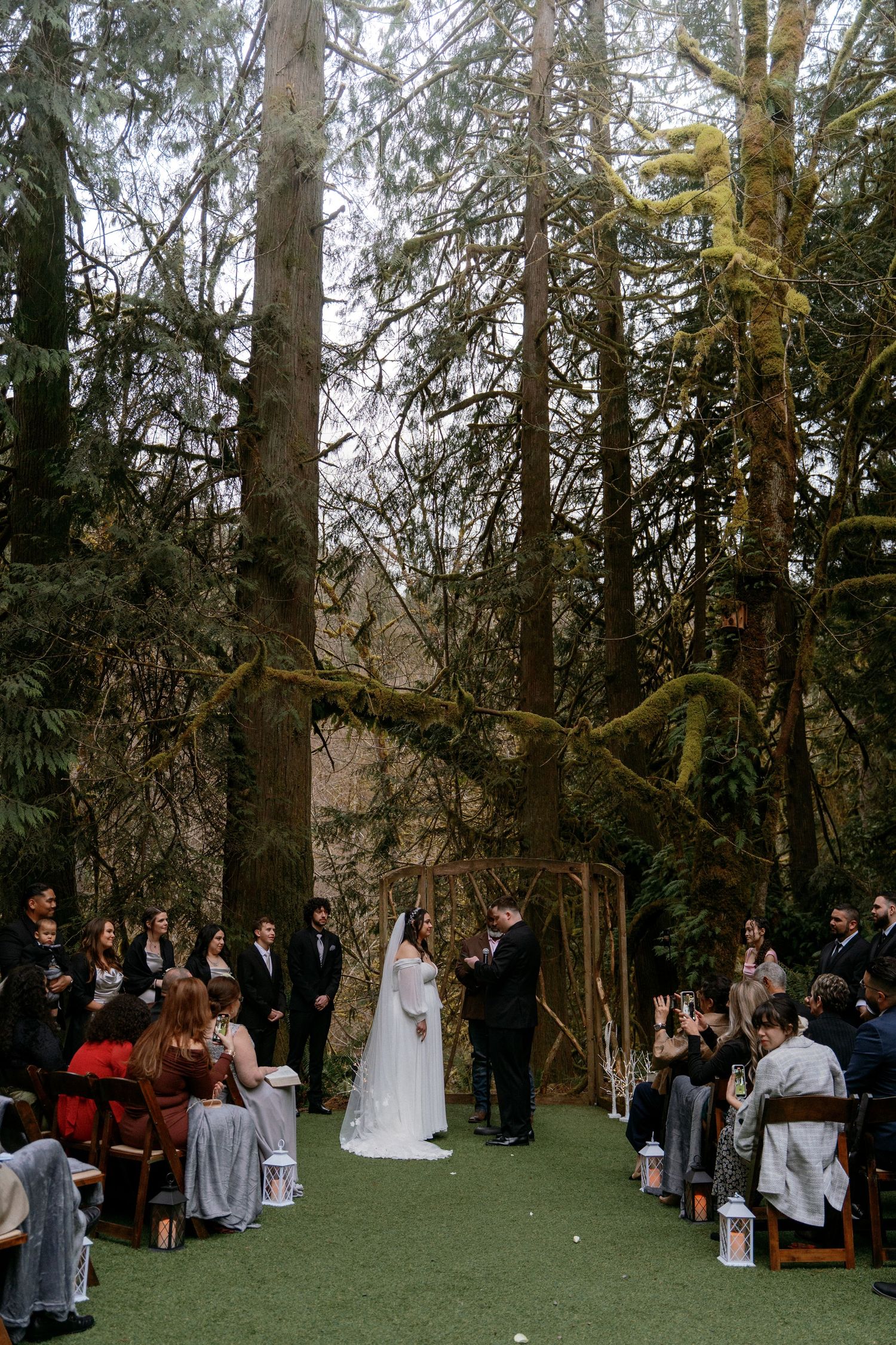 Outdoor forest wedding ceremony with bride and groom at rustic wooden arch surrounded by tall trees.