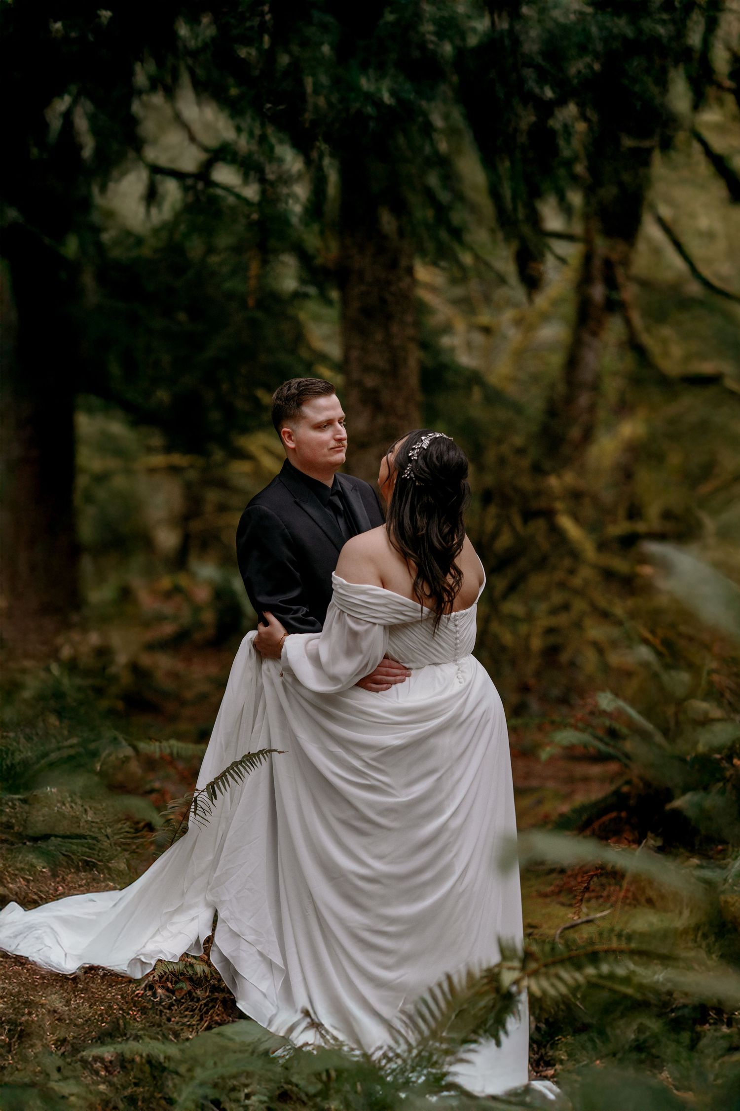 Groom embracing bride in flowing white dress while dancing among ferns in a lush forest wedding.