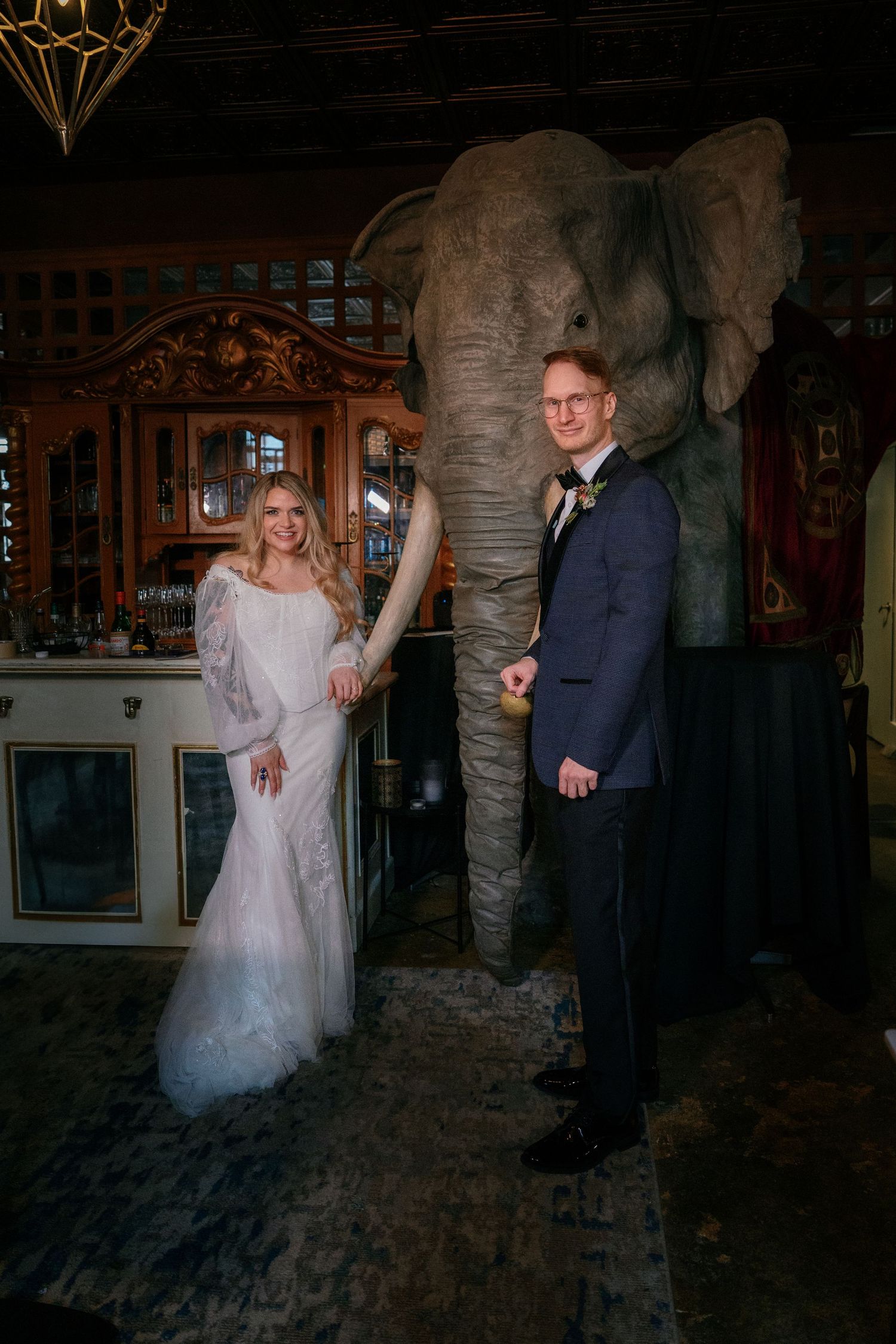 Smiling bride and groom pose holding hands in front of large elephant sculpture in an ornate vintage venue interior.