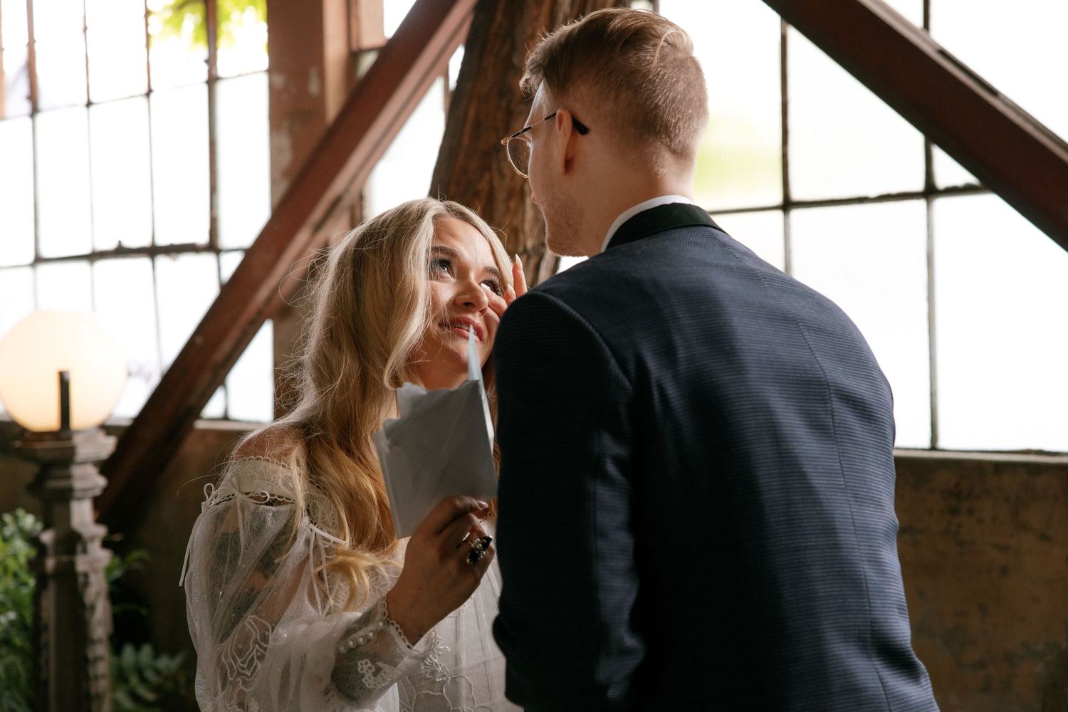 Bride and groom share an intimate moment during wedding ceremony in rustic industrial venue with large windows.