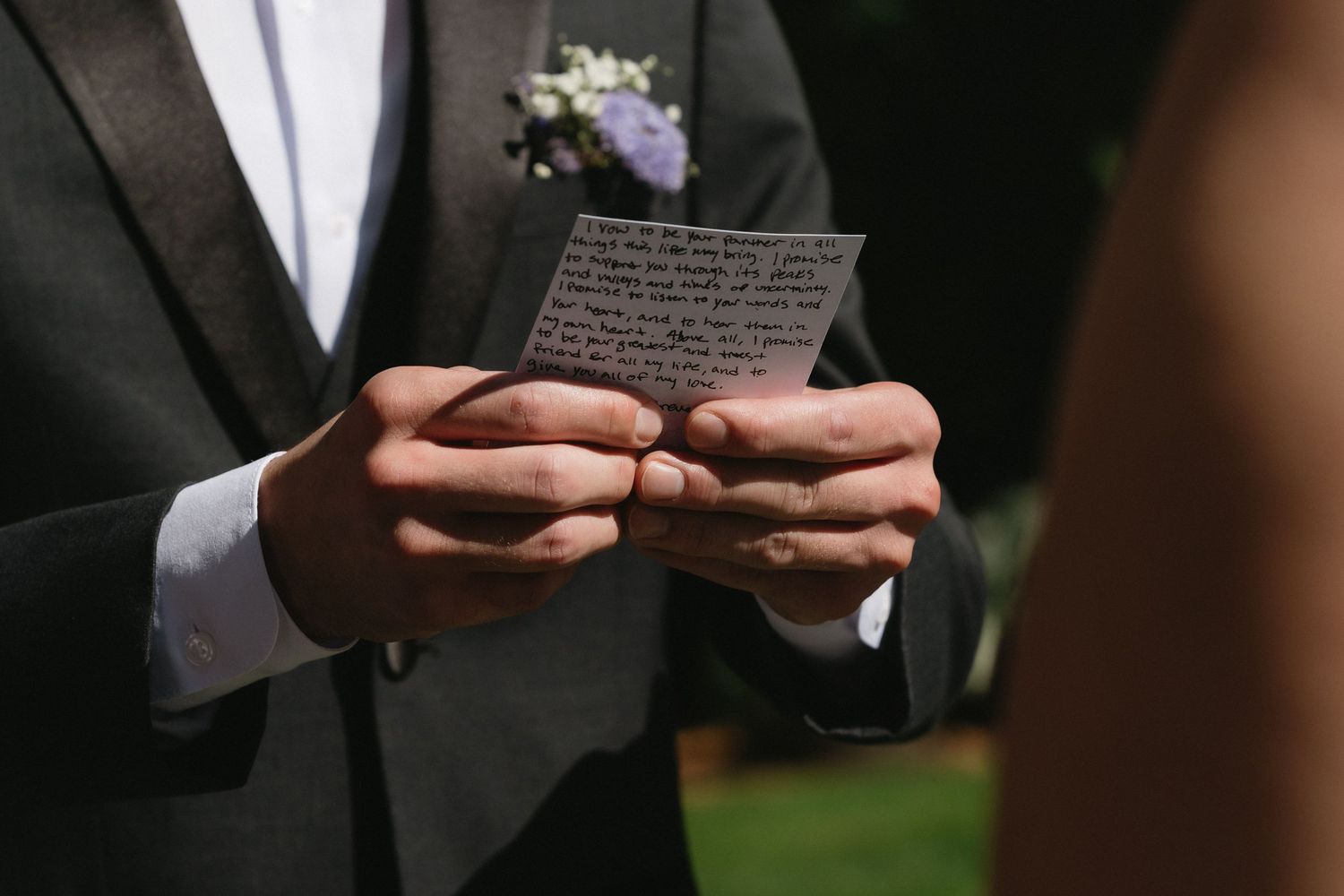 Groom in gray suit holds handwritten wedding vows card during outdoor ceremony with boutonniere on lapel.