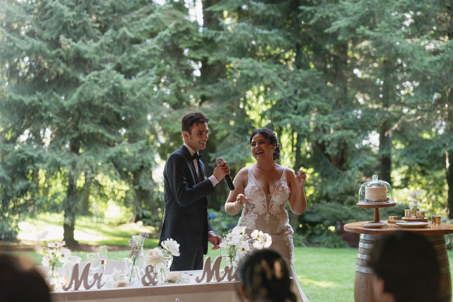 Bride in lace gown and groom in suit clapping at outdoor wedding reception sweetheart table.