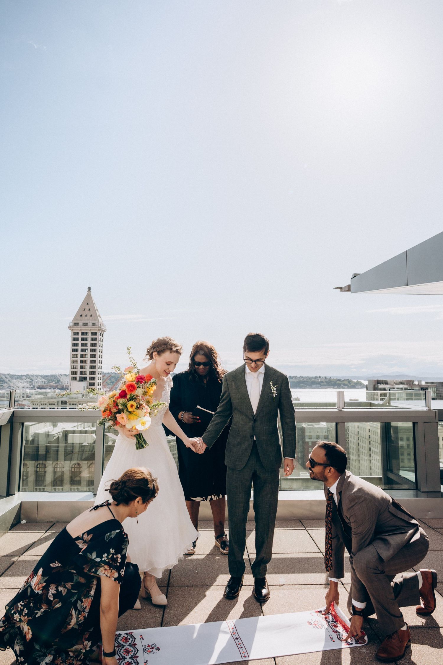 Bride and groom sign wedding certificate on sunny rooftop with city skyline and waterfront views.