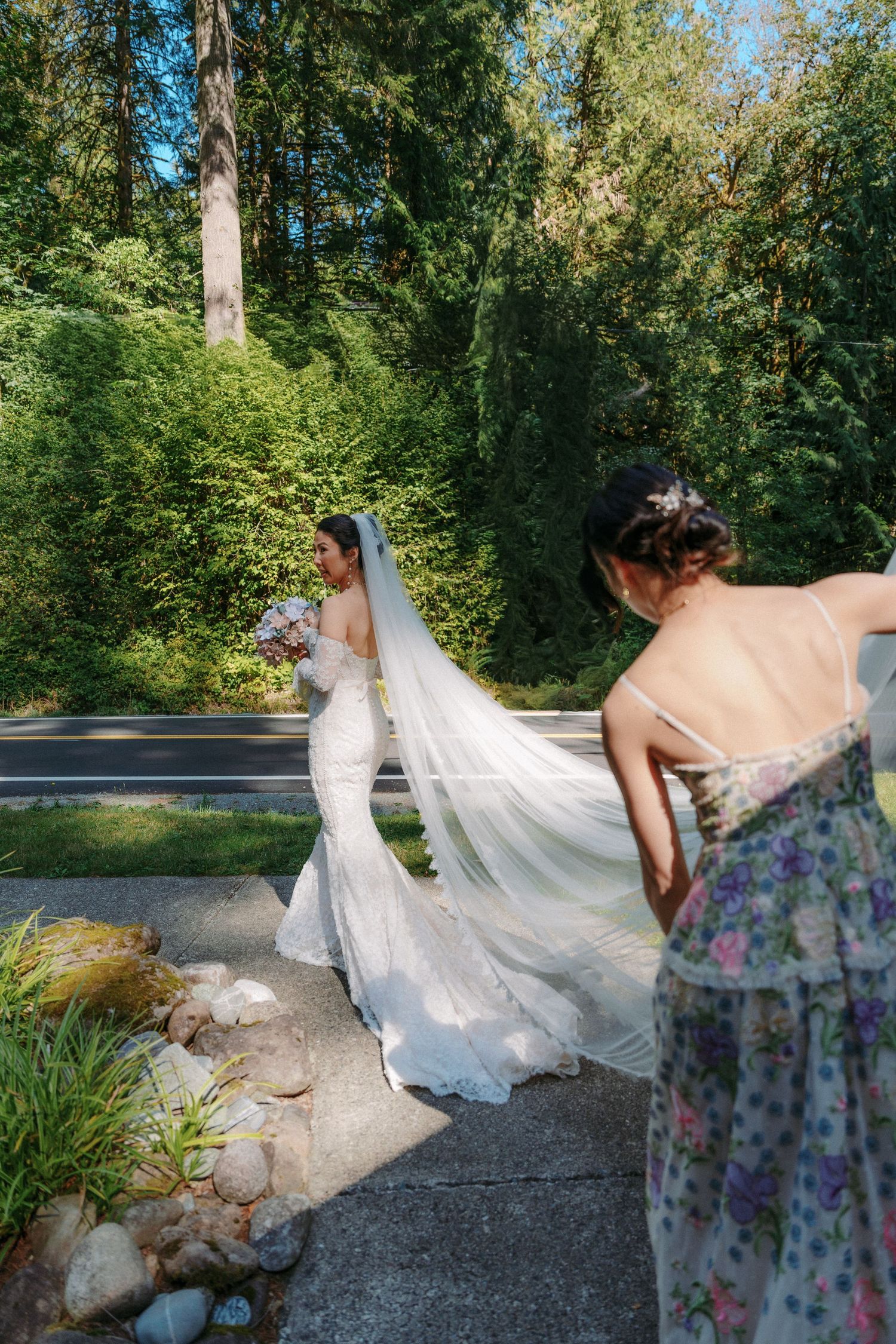 Bride in elegant white gown and veil walks outdoors as bridesmaid adjusts her train in forest setting.