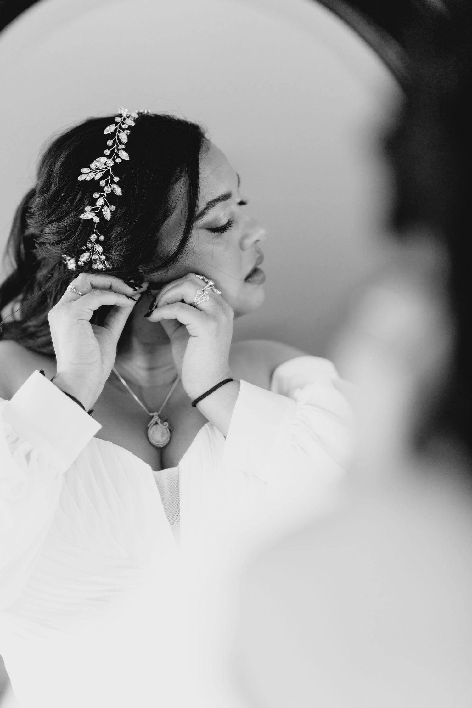 Black and white photo of bride putting on earrings in mirror, wearing floral hair vine and robe.