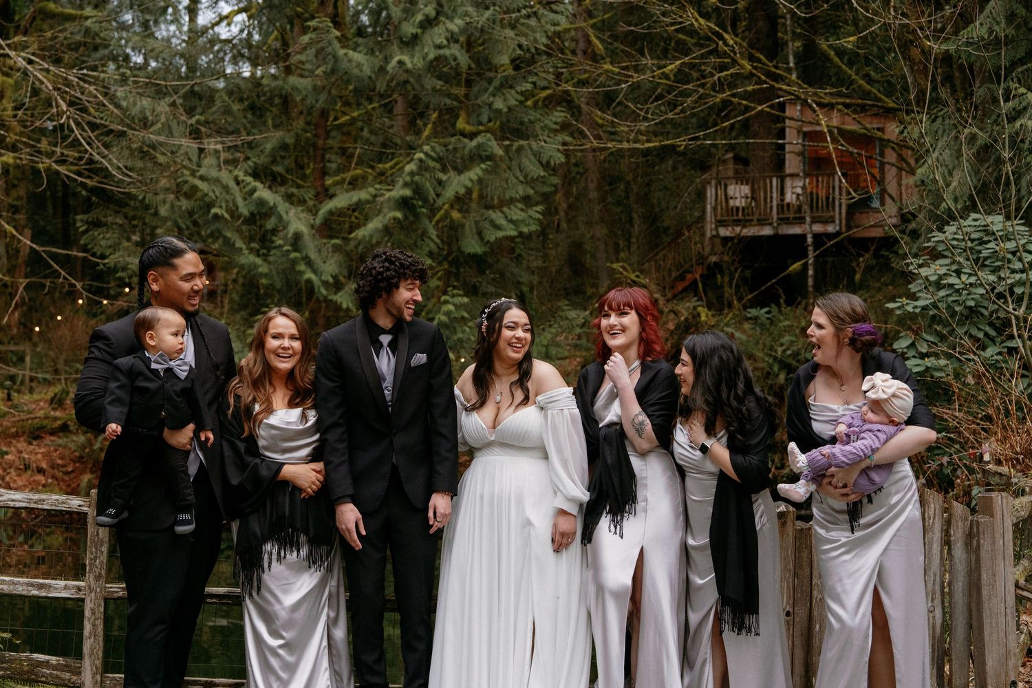 Wedding party group laughing outdoors in forest, bride in white gown flanked by bridesmaids in silver dresses.