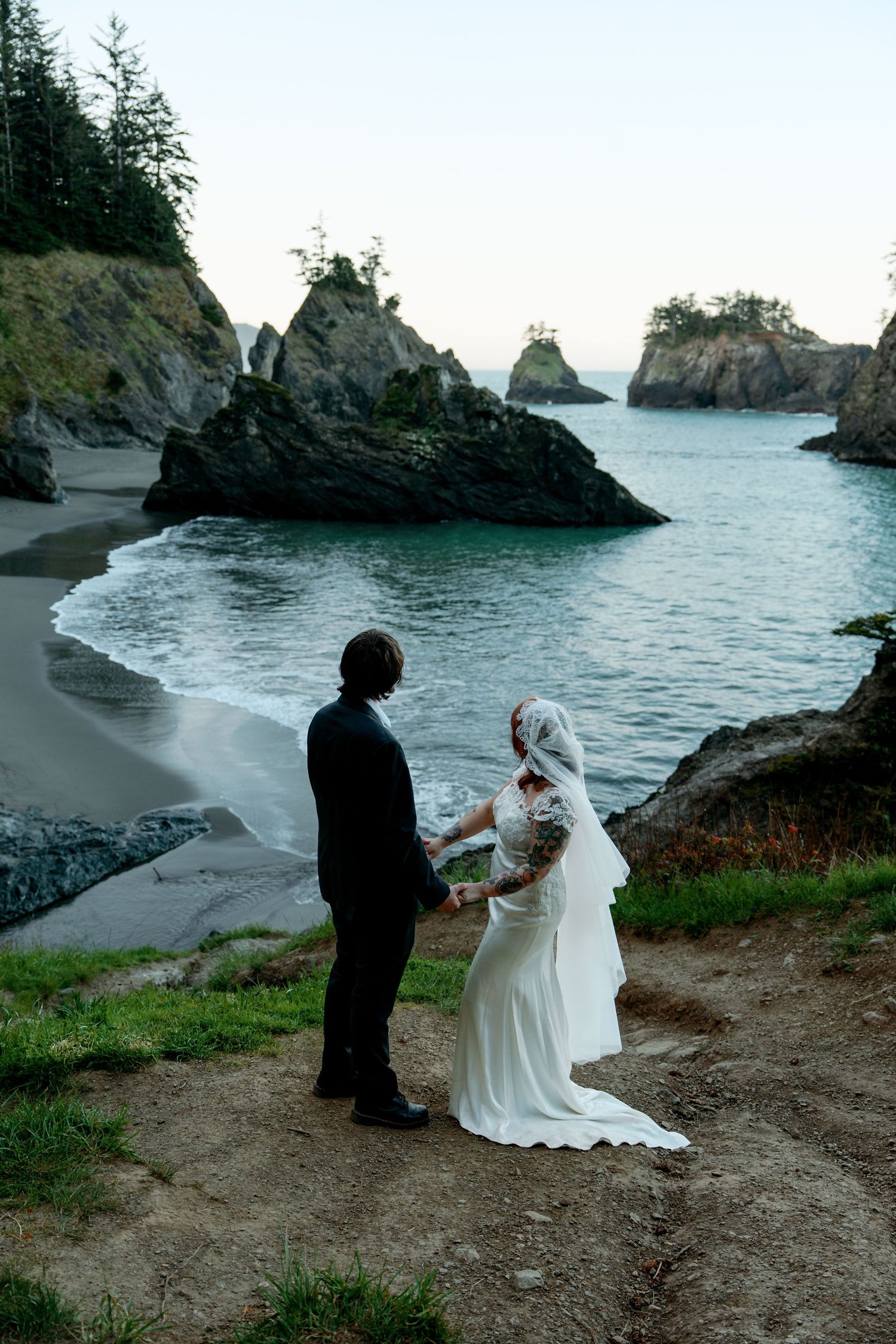 Bride and groom holding hands on a cliff overlooking a stunning rocky ocean cove at dusk.