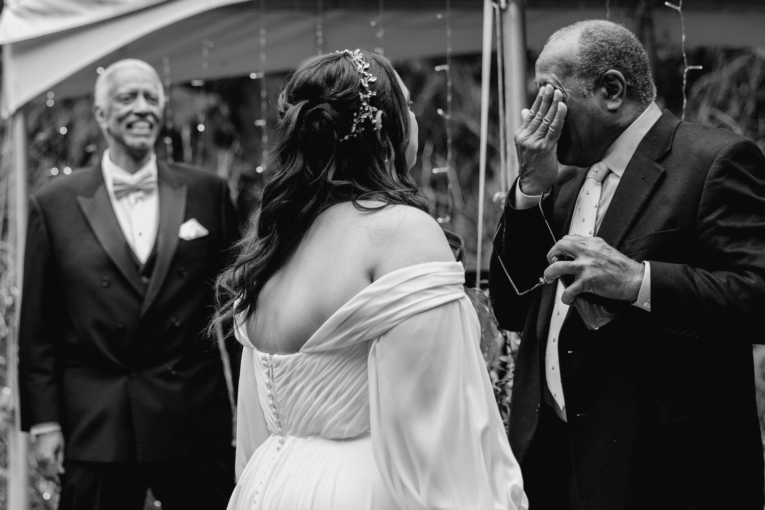 Emotional father wipes tears seeing bride at wedding ceremony in black and white photo.