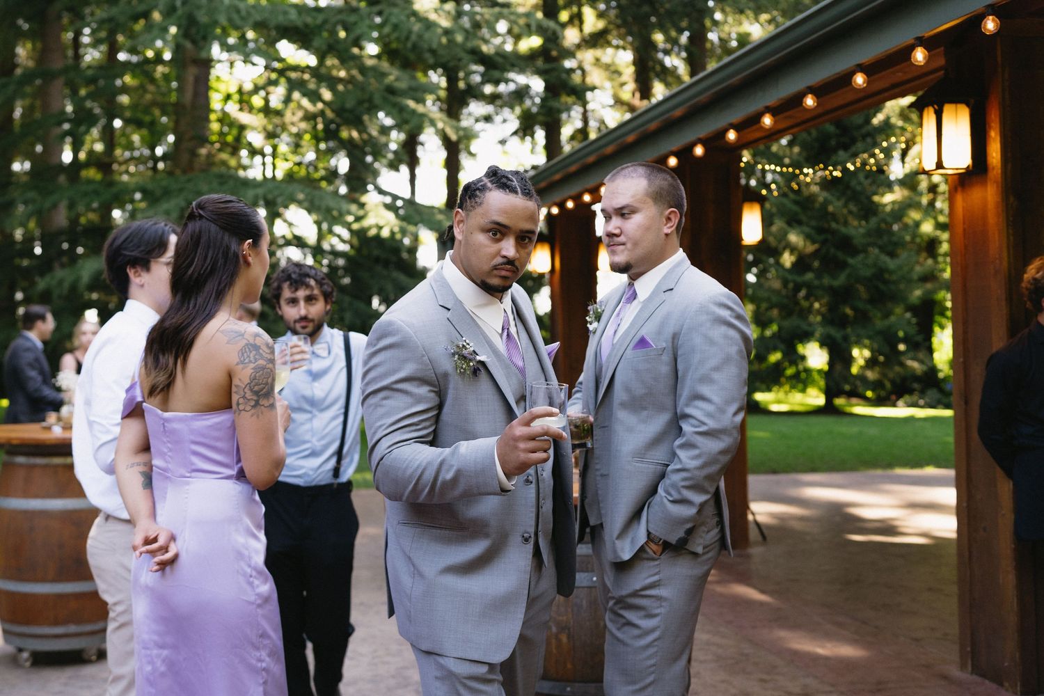 Two groomsmen in gray suits chat at outdoor rustic wedding reception venue with string lights.