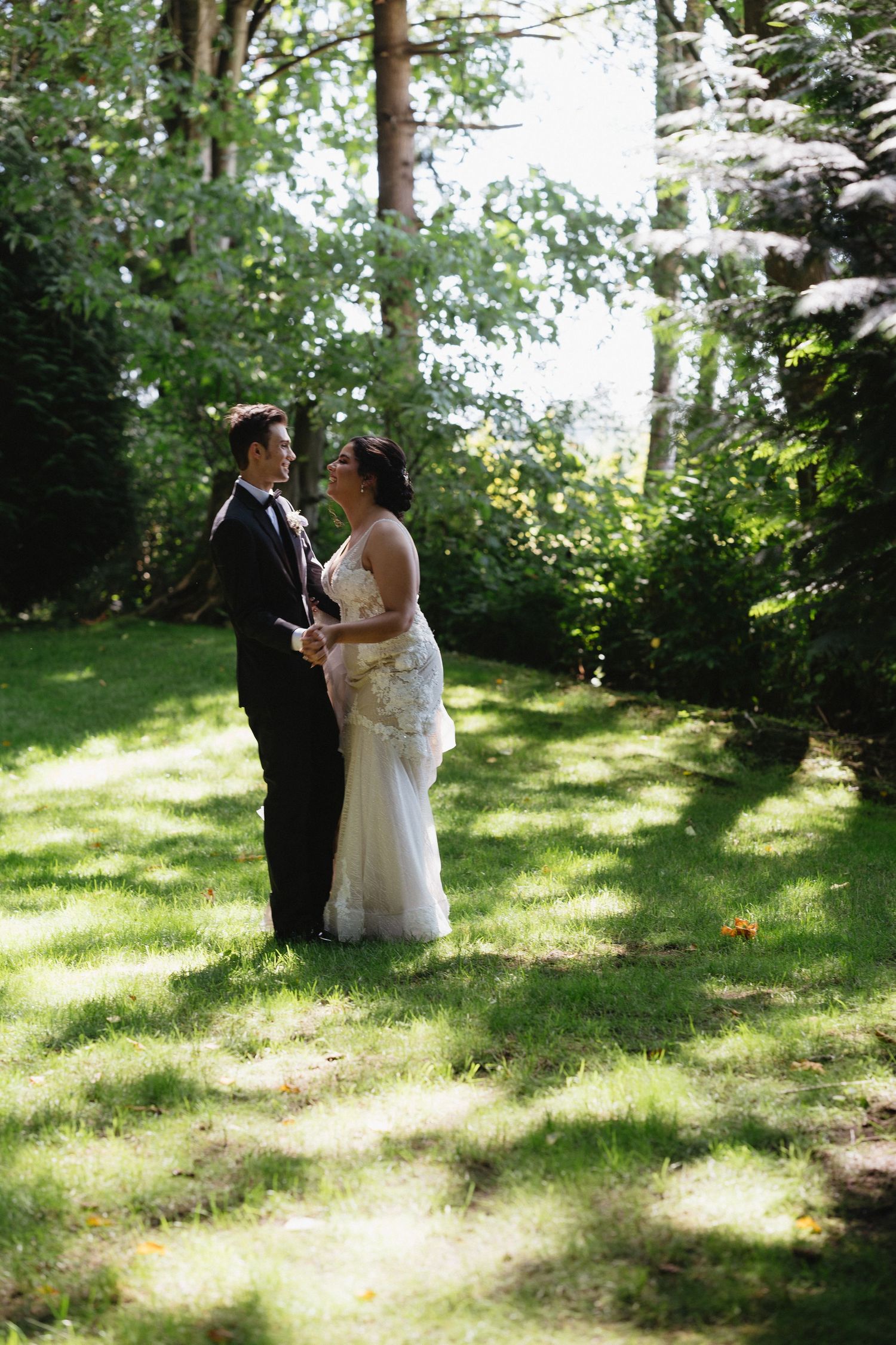 Bride in lace gown and groom in black tuxedo standing together in a sunny tree-lined garden.