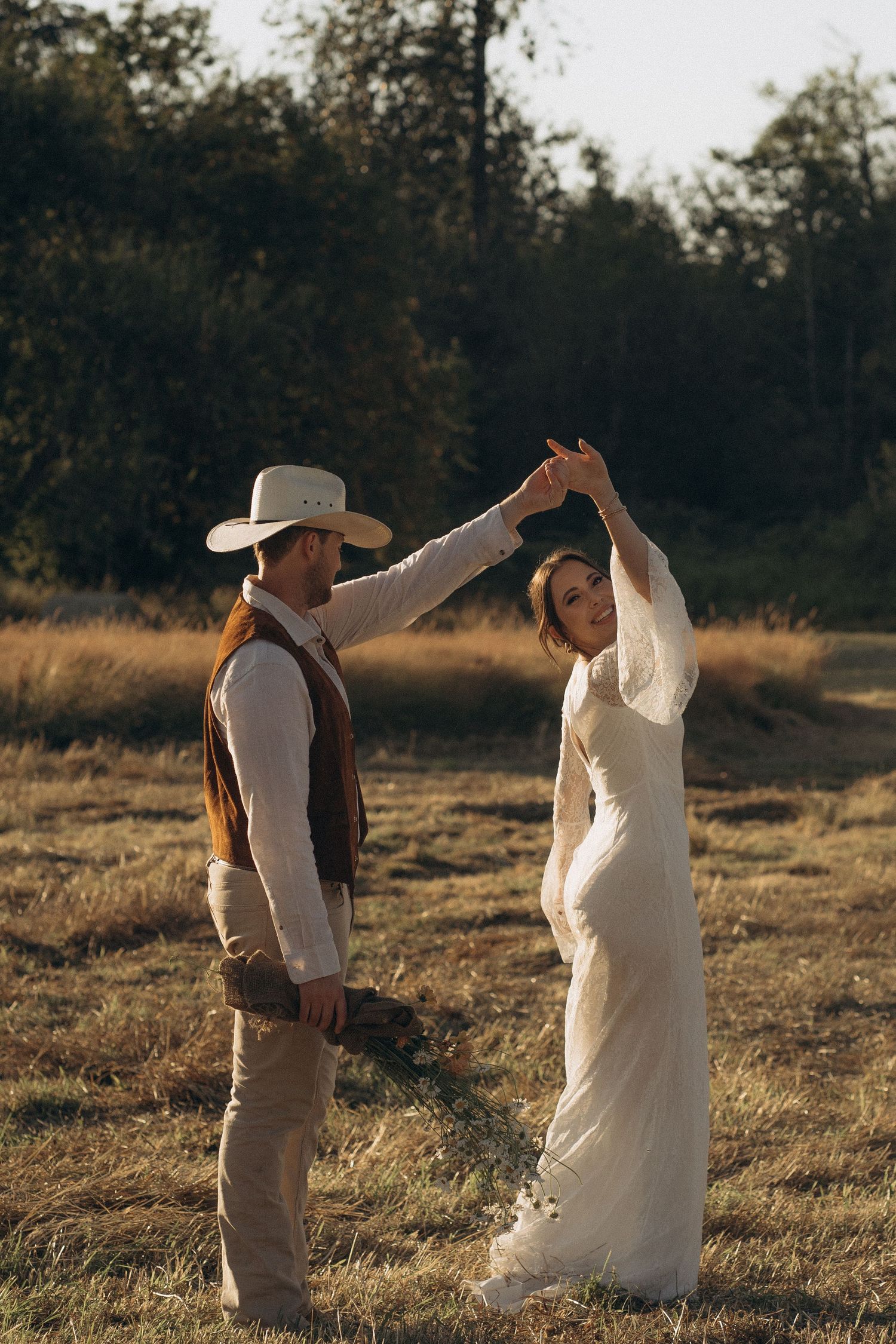 Groom in cowboy hat spinning bride in white dress during outdoor rustic wedding shoot.