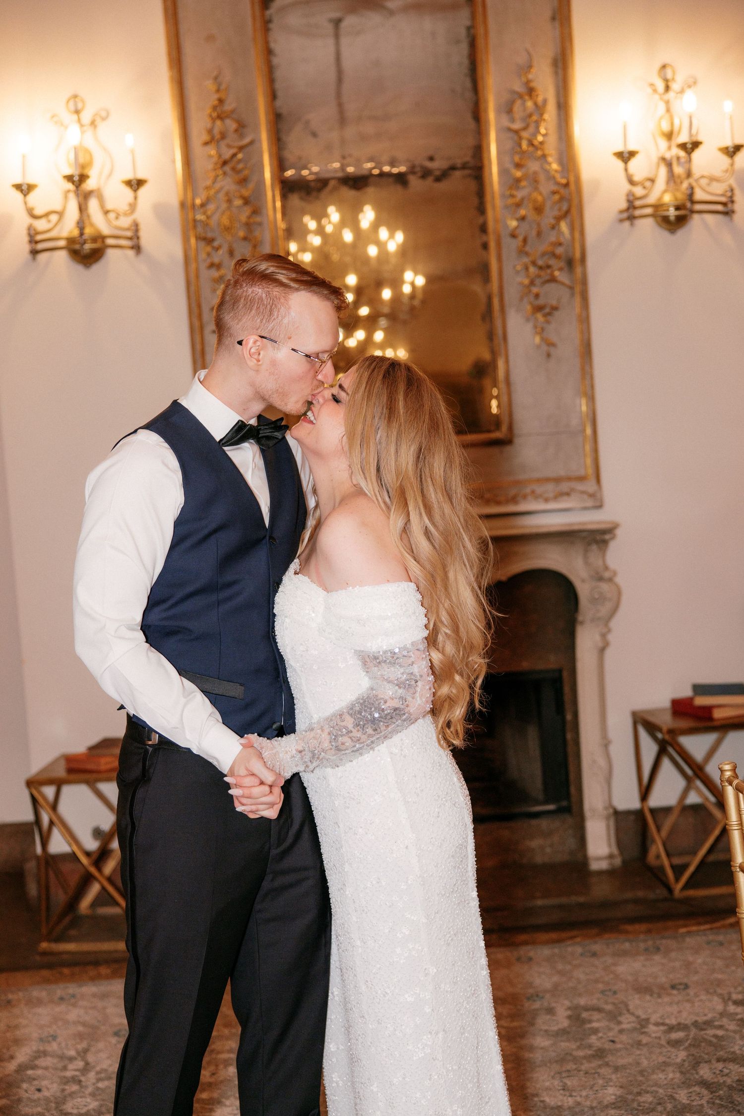 Bride and groom sharing a kiss in an ornate ballroom with golden mirror and glowing wall sconces.