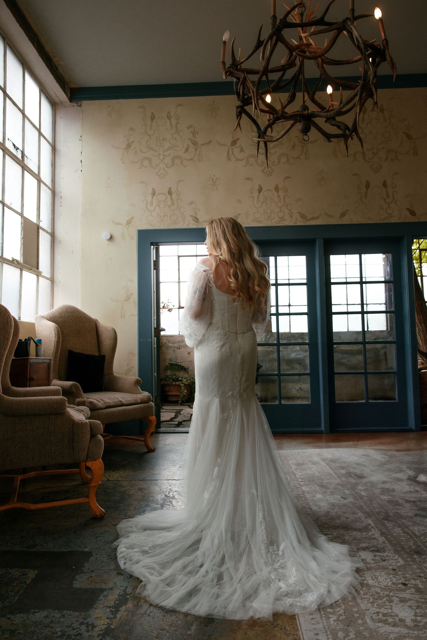 Bride in lace mermaid gown with train stands before blue French doors under antler chandelier.