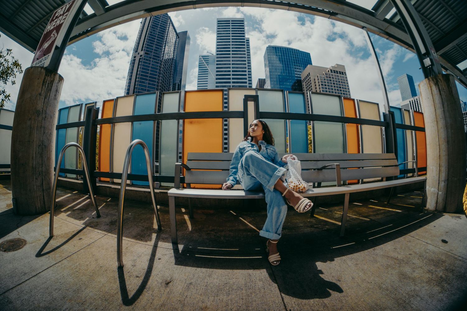 Woman in denim outfit sits on a bench with city skyscrapers and colorful panels behind her, fisheye lens shot.