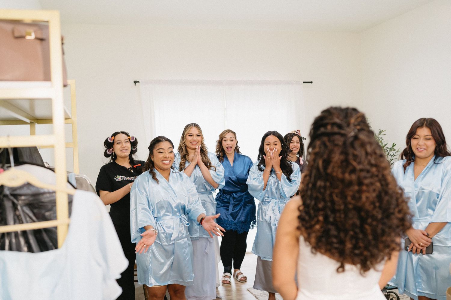 Bridesmaids in blue satin robes reacting with joy as bride reveals wedding dress.
