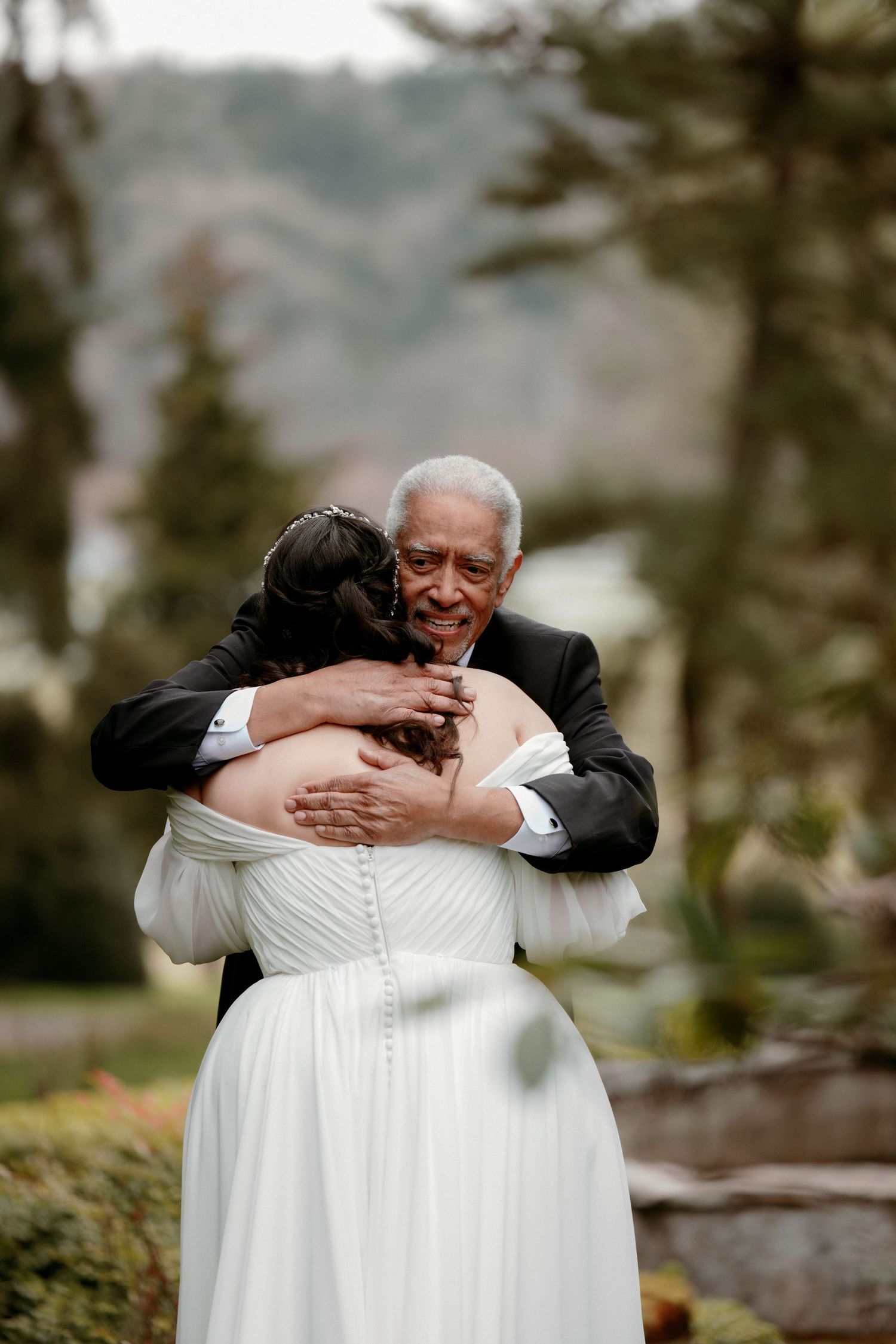 Elderly man in suit shares a warm embrace with bride in white off-shoulder gown at outdoor wedding.