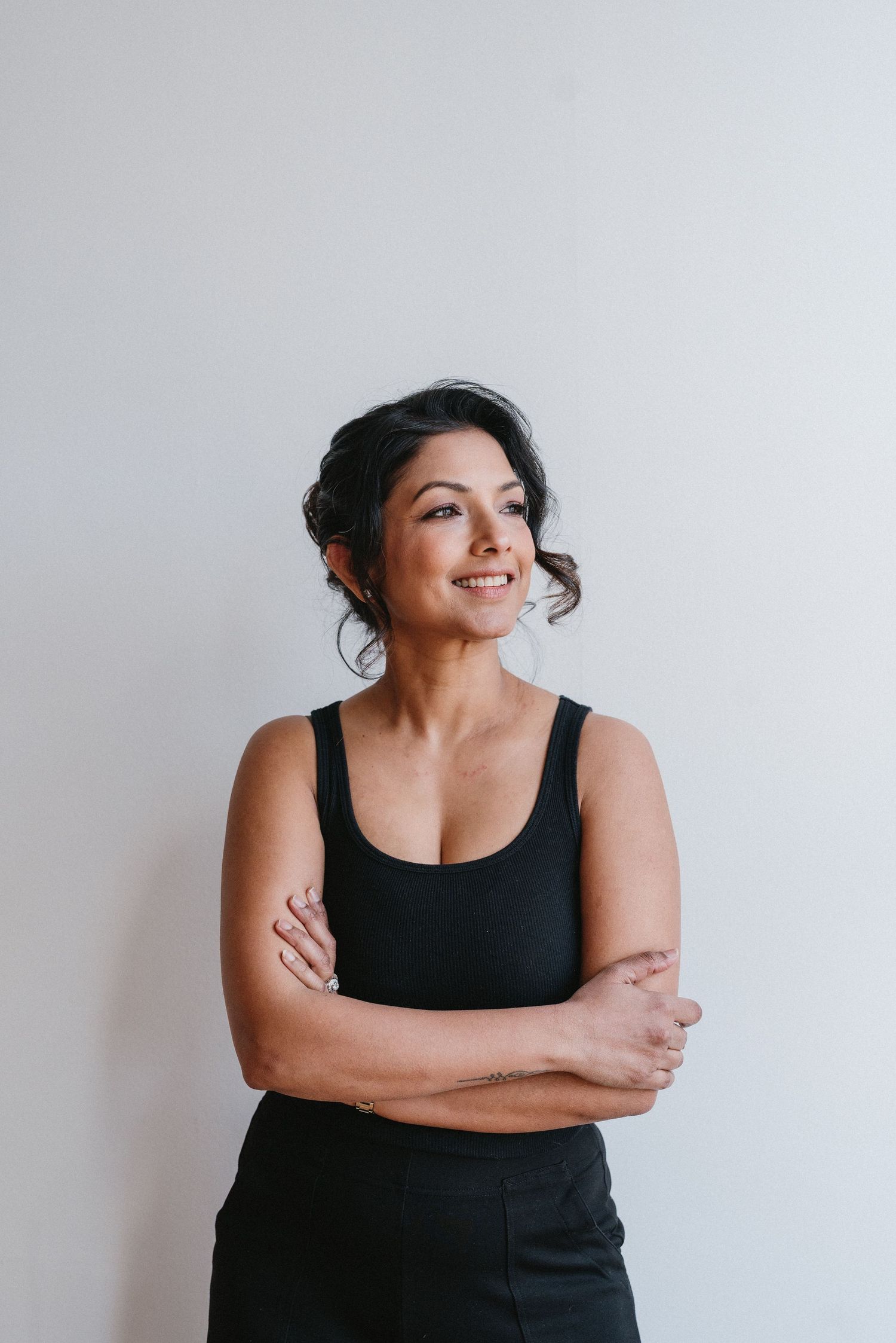 Smiling dark-haired woman in black tank top poses with arms crossed against white wall.