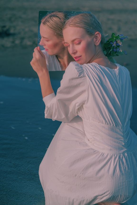 Blonde woman in white dress holding mirror reflecting her face at beach at dusk.