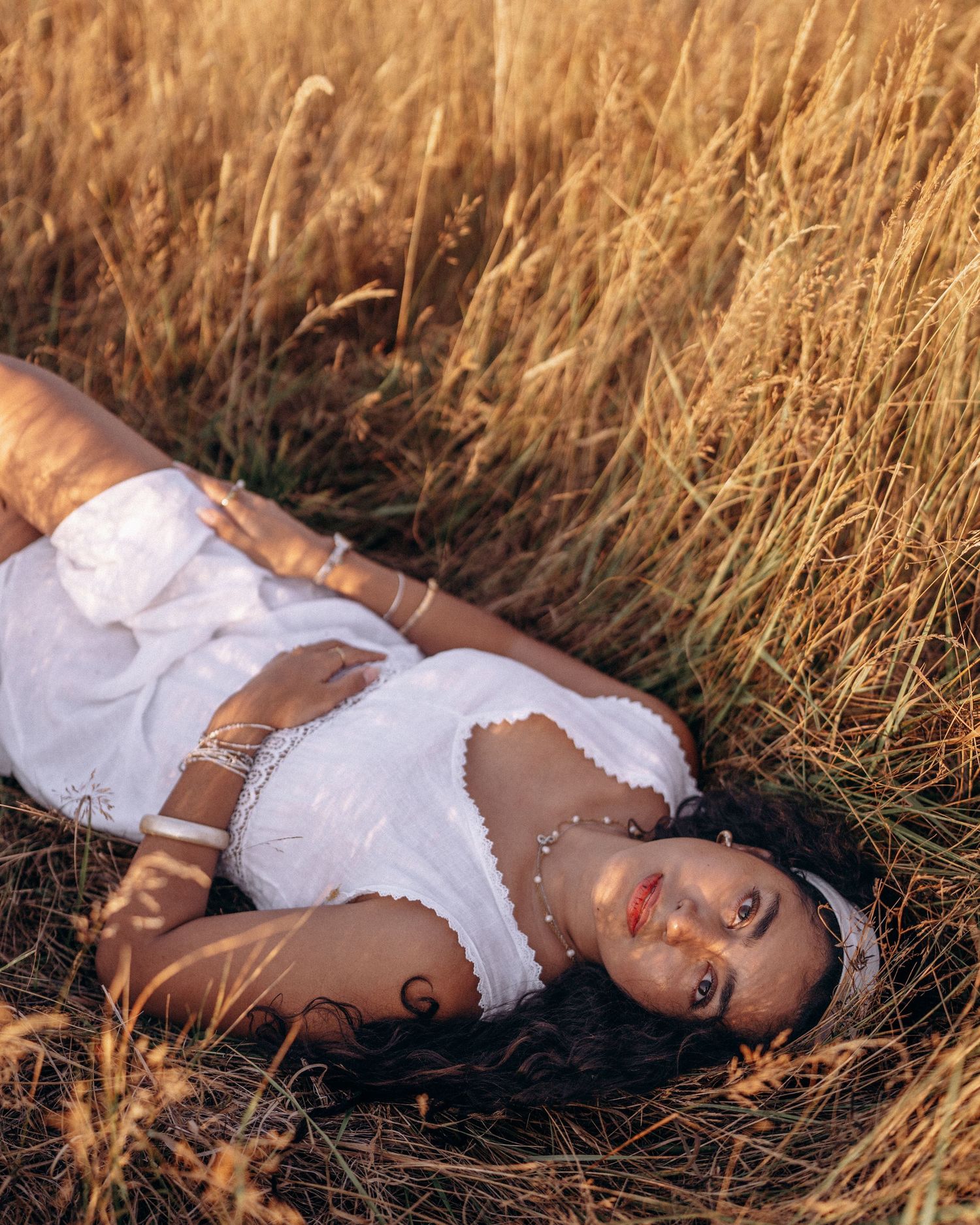 Woman in white lace dress lying in tall golden grass field, wearing silver jewelry and headband.