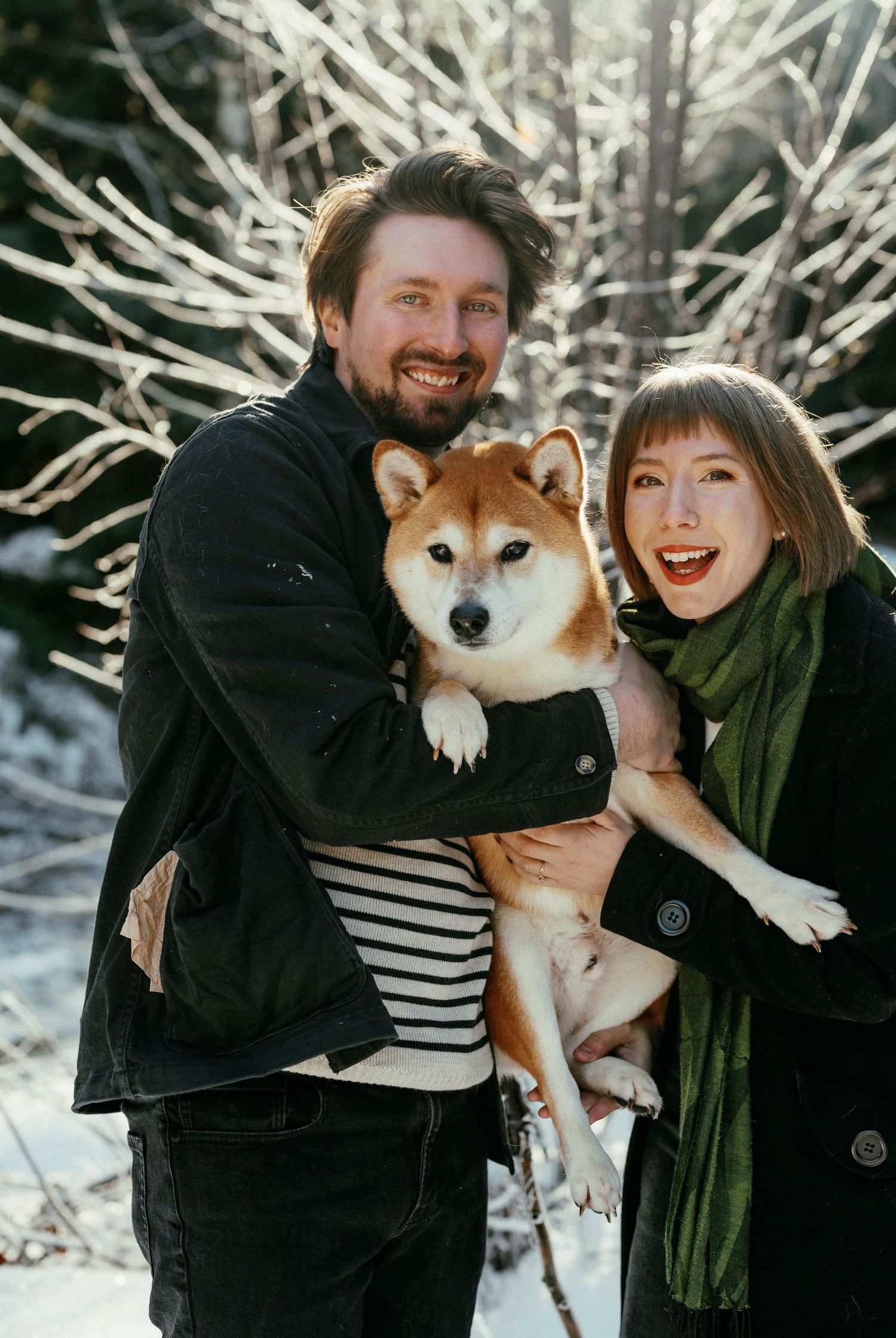 Smiling couple holding a Shiba Inu dog in a snowy winter forest with sunlit bare branches.