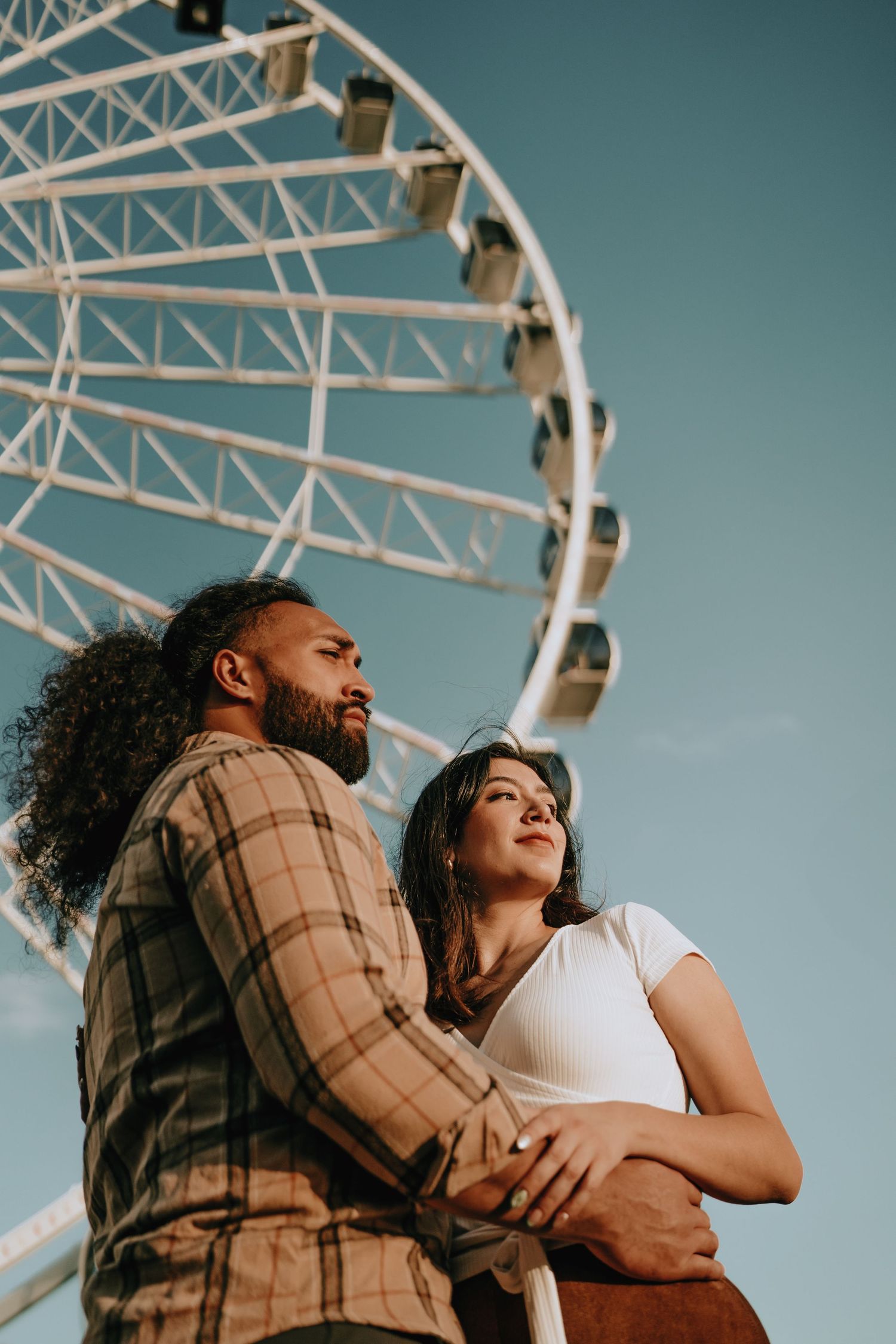 Romantic couple standing together in front of a large white Ferris wheel under a clear blue sky.