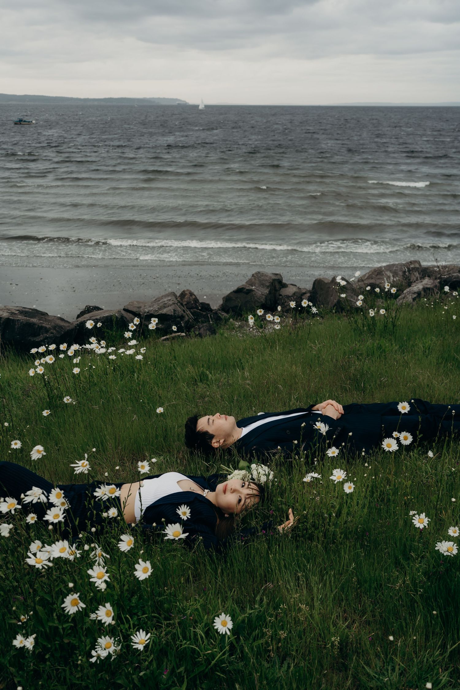 Couple in dark suits lying in field of white daisies beside rocky ocean shoreline.