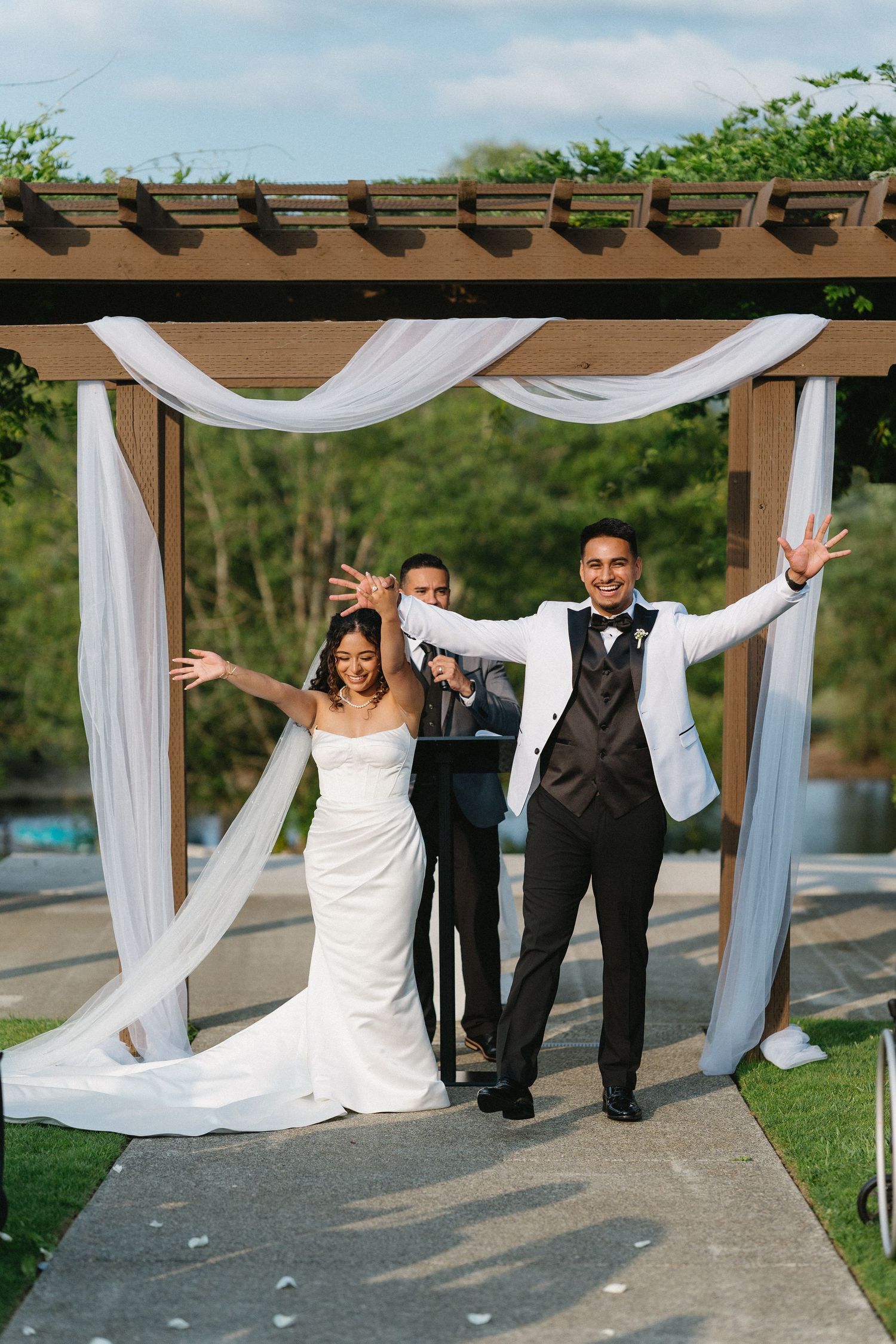 Joyful newlyweds celebrate with raised arms under white-draped pergola after ceremony.