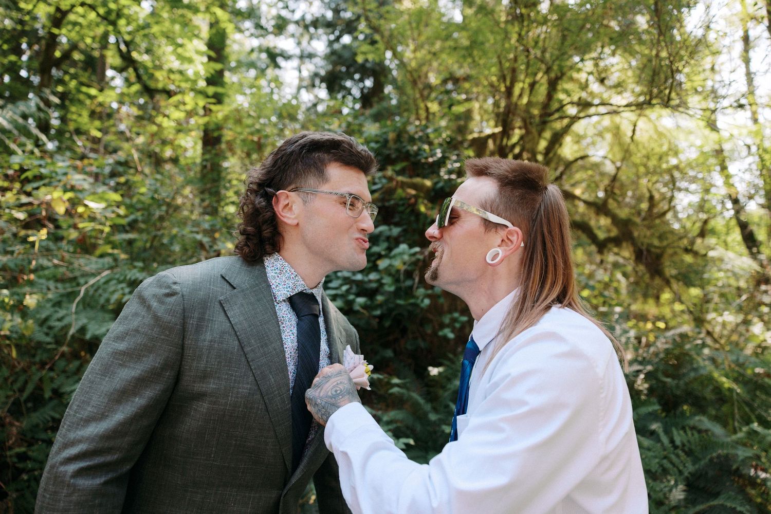 Two men in suits share a playful moment together in a lush green forest setting.