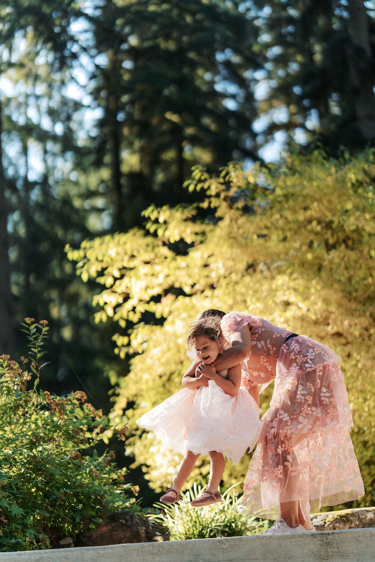 Woman in floral gown helps a little girl in white tutu dress near garden greenery in golden sunlight.