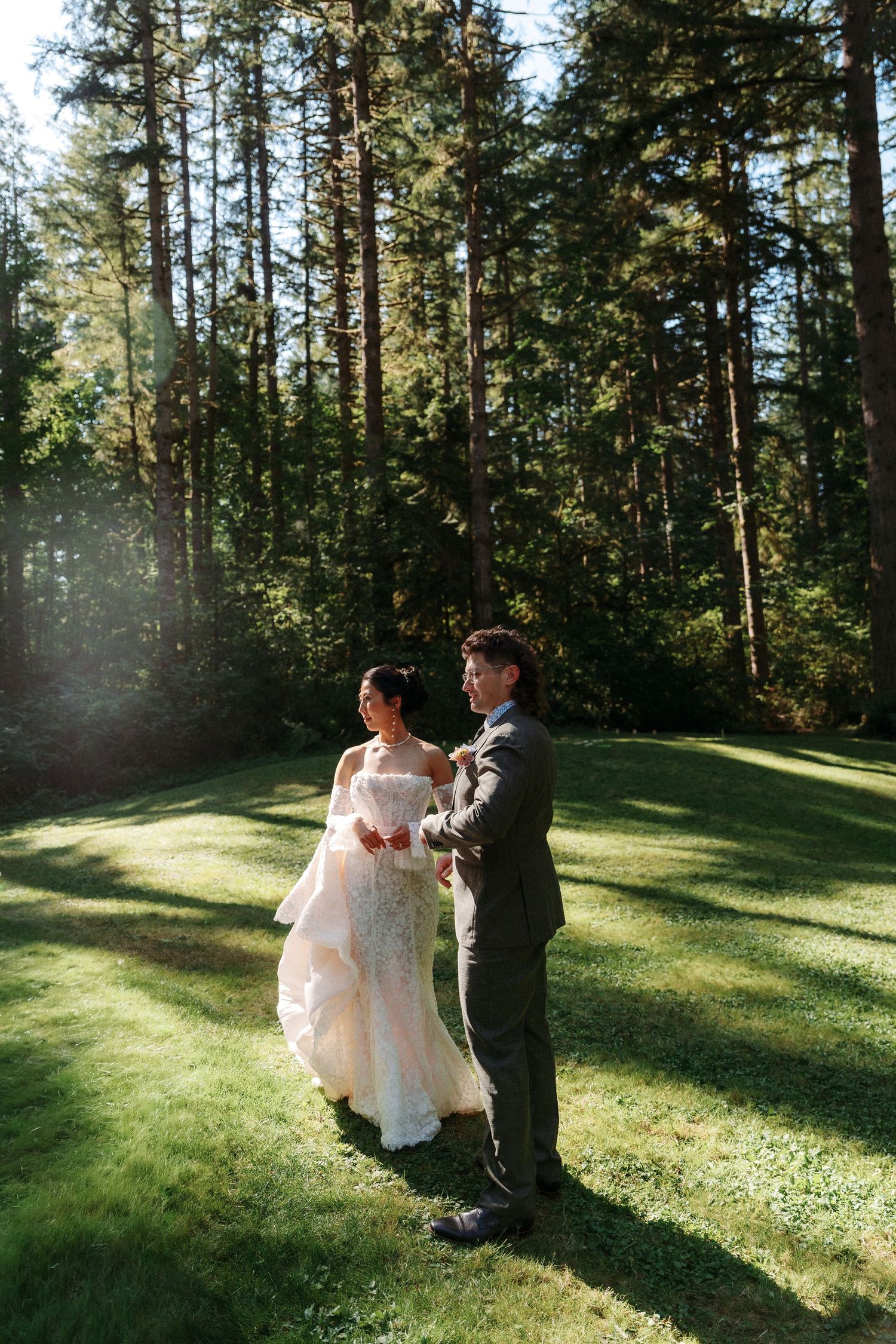 Bride and groom dance together on sunlit green lawn surrounded by tall pine trees.