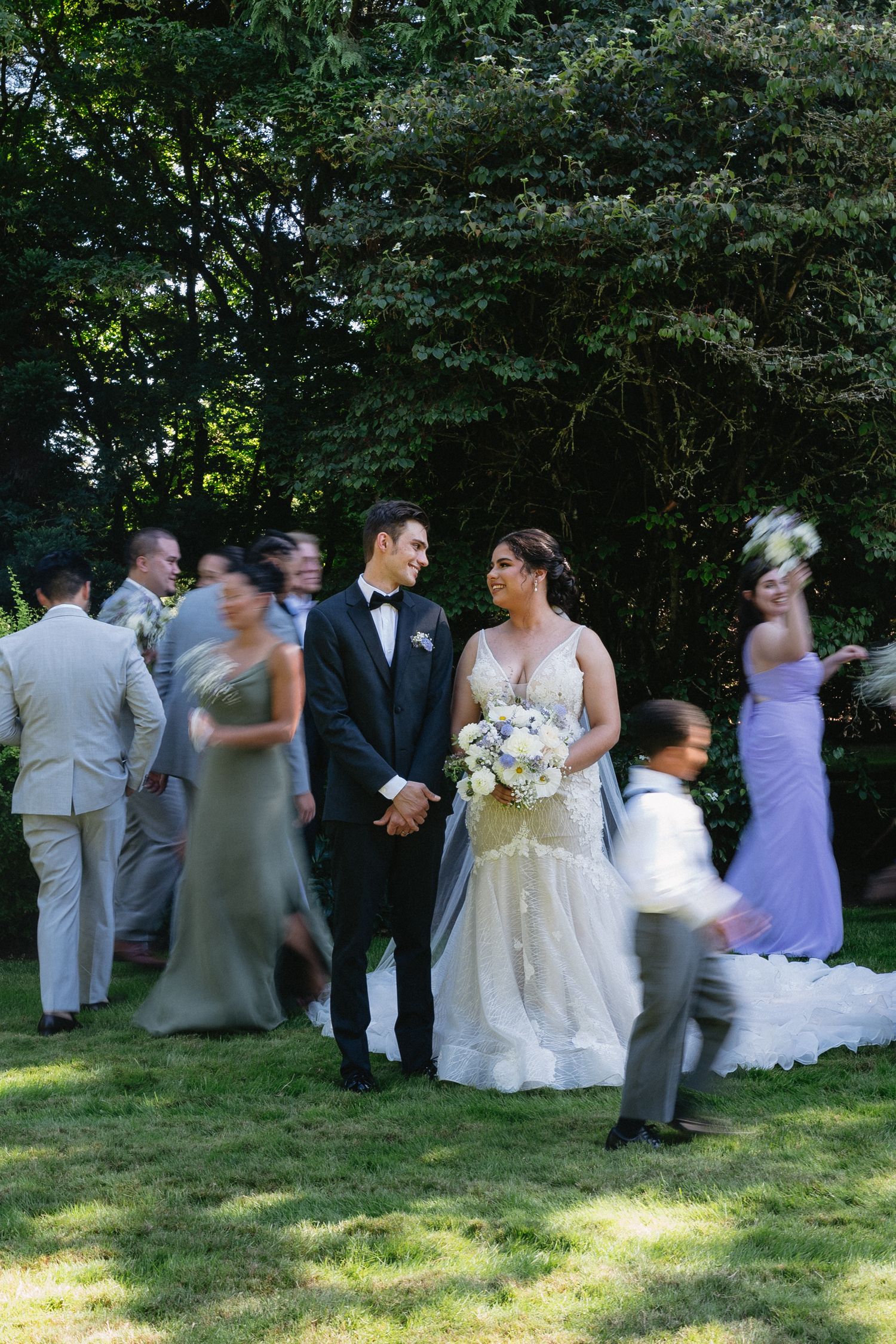 Bride holding white bouquet and groom in tuxedo stand with blurred wedding party on lawn.
