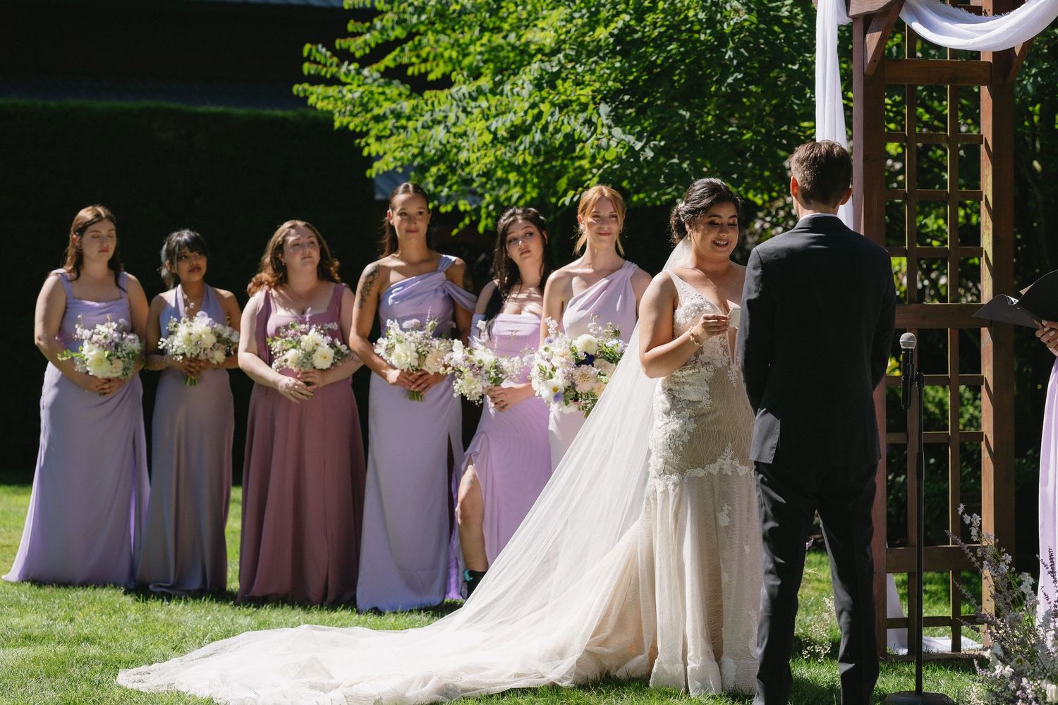 Bride in lace gown faces groom at altar with bridesmaids in lavender and mauve dresses holding bouquets.