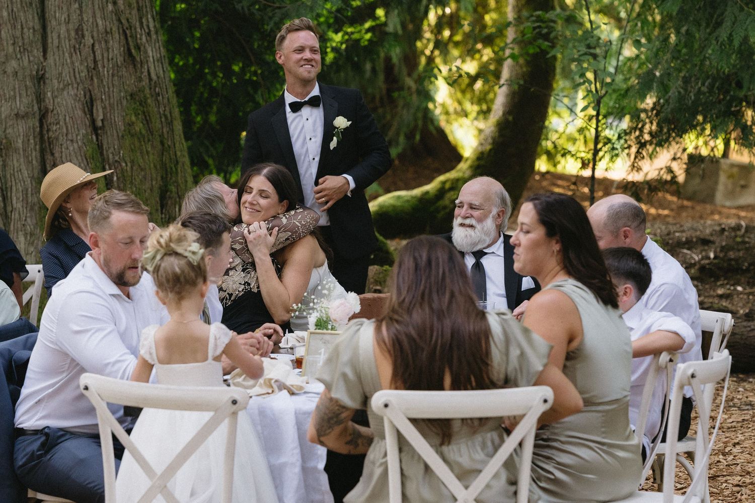 Wedding guests gathered at outdoor reception tables under trees, sharing joyful moments during speeches.