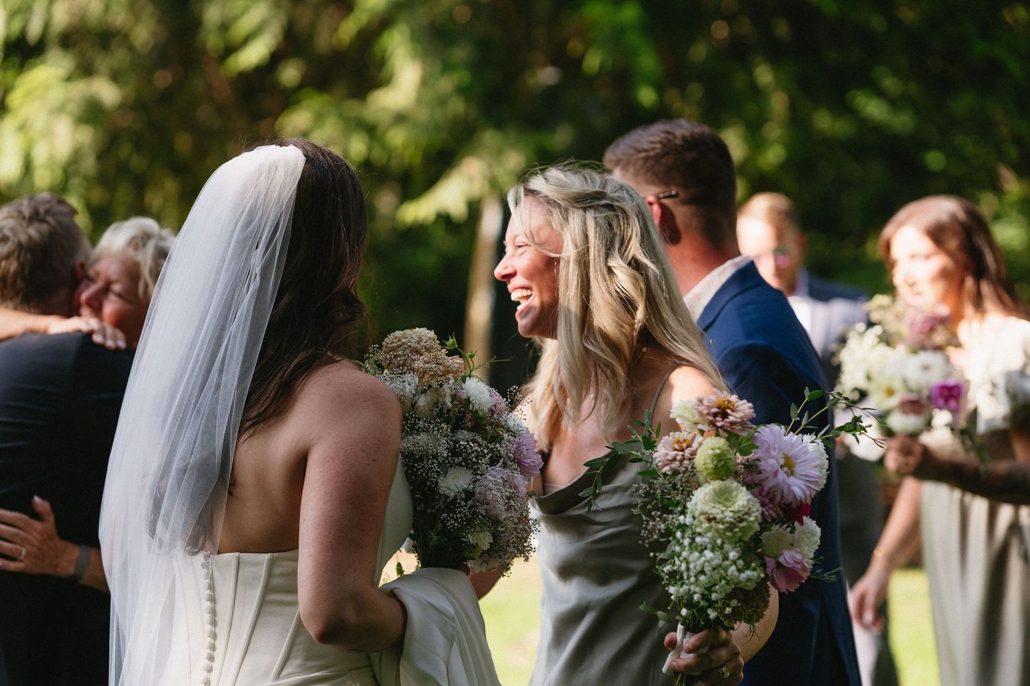 Bride in white veil embracing smiling bridesmaid holding floral bouquet after outdoor wedding ceremony.