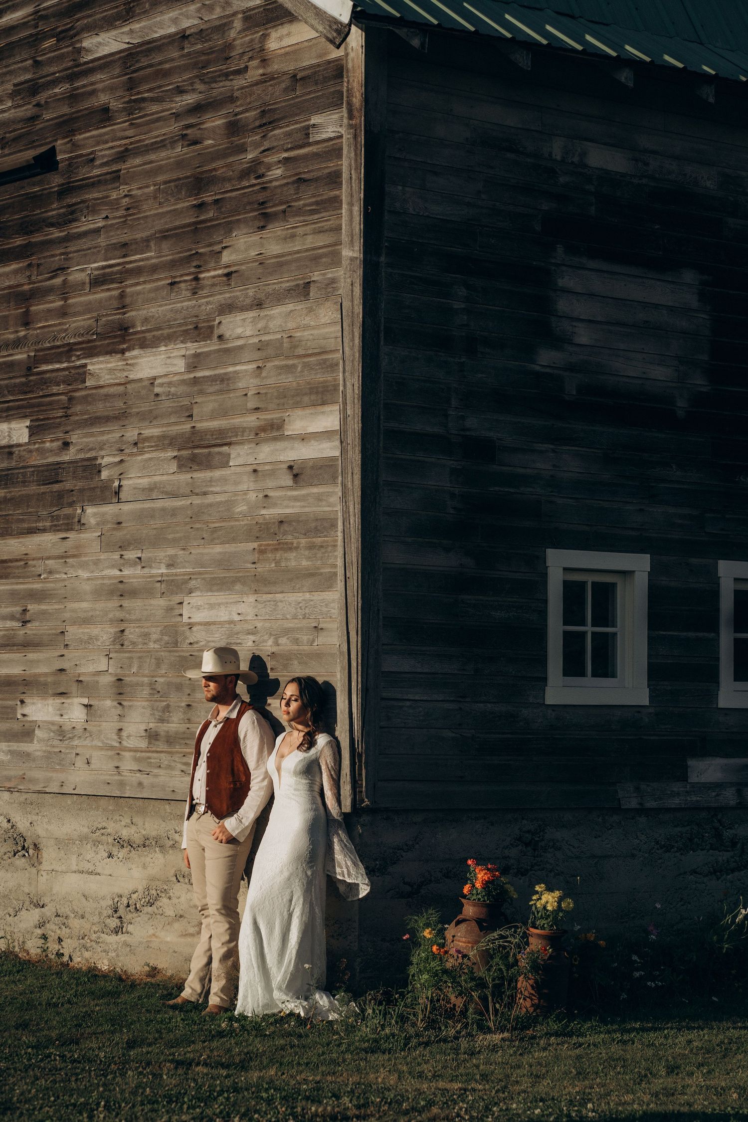 Bride in white lace gown and groom in cowboy hat lean against a weathered wooden barn at sunset.